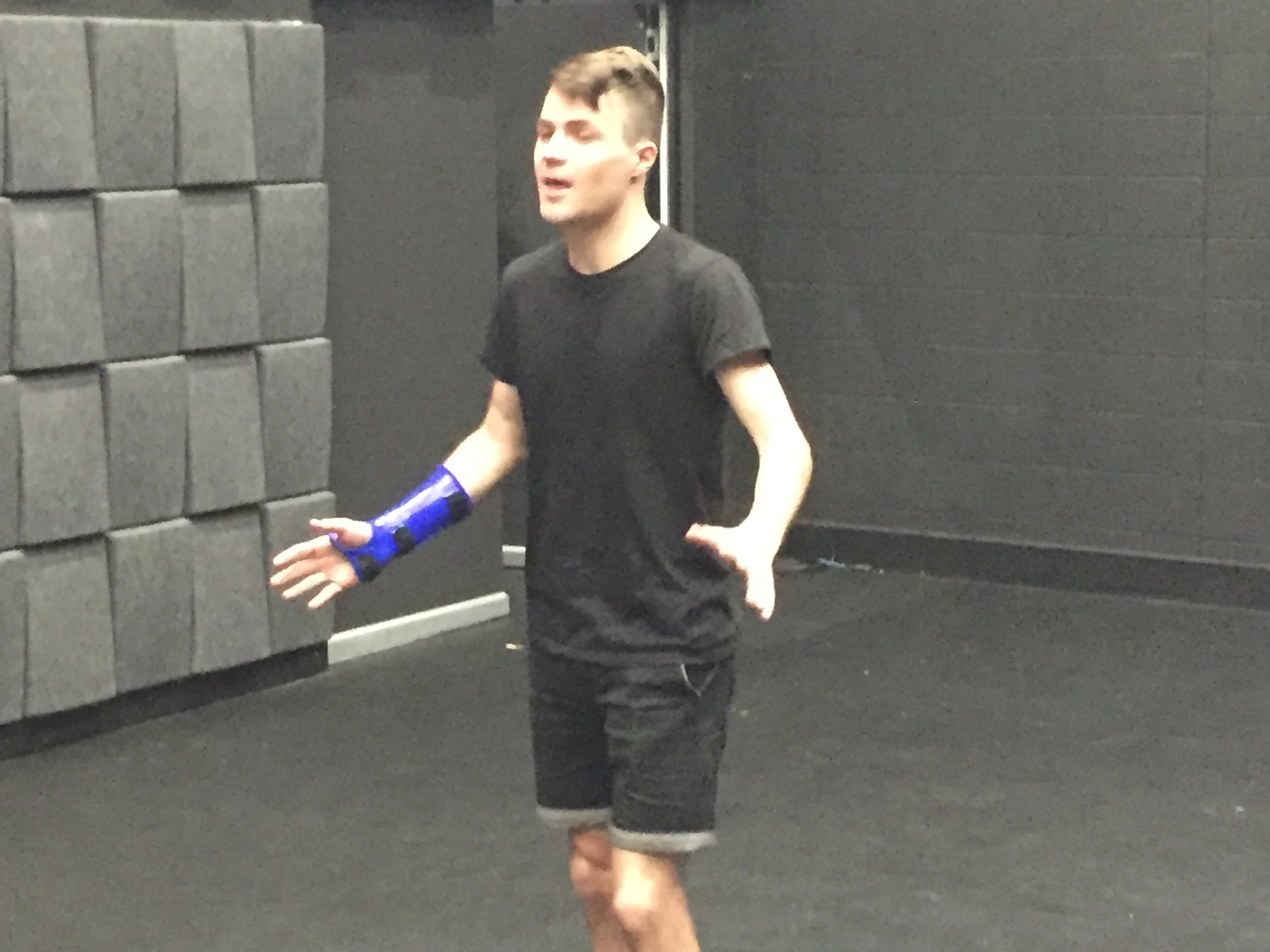 Man wearing a black T-shirt and shorts, gesturing with a blue wrist brace in an indoor gym.