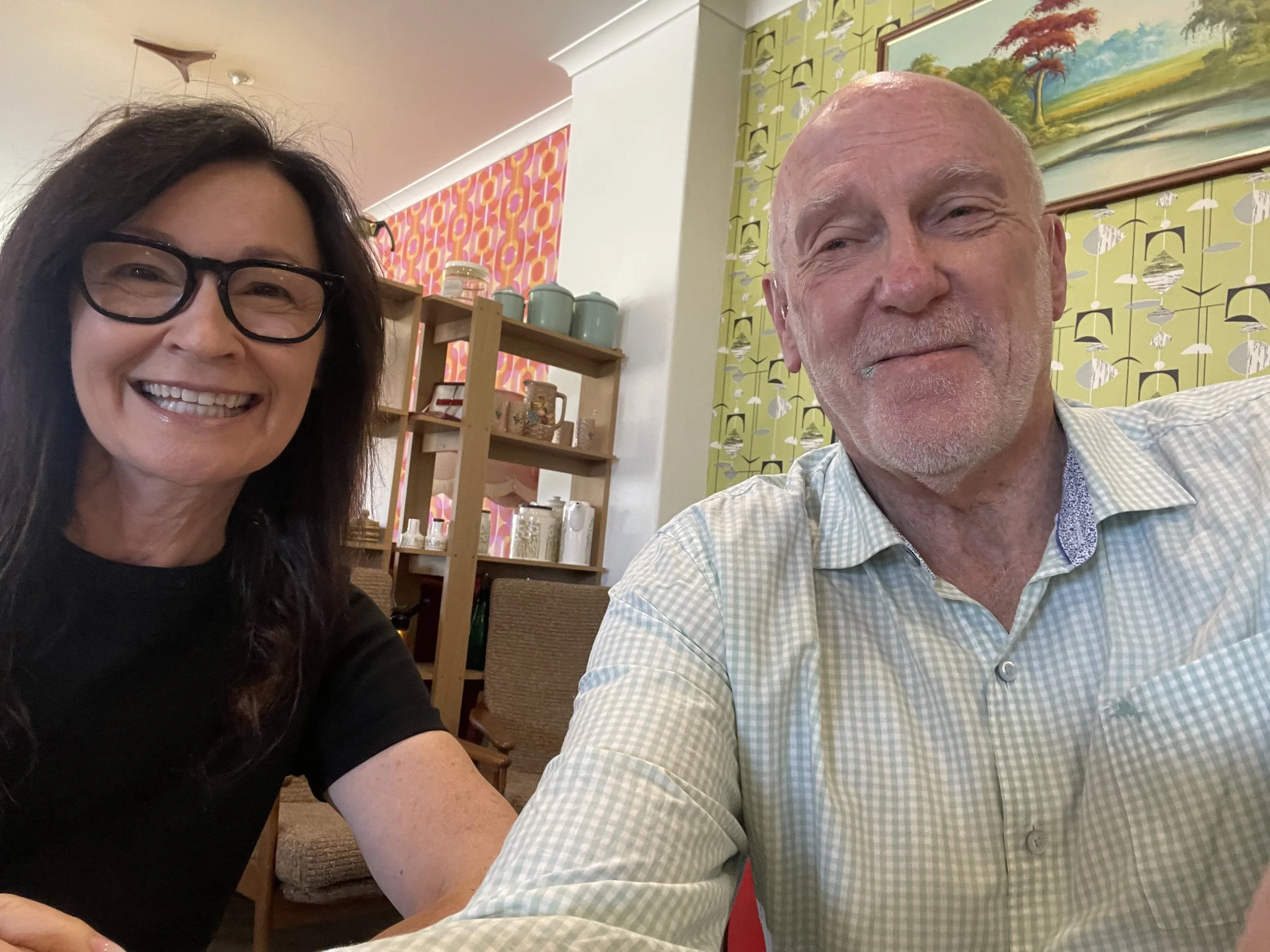 Smiling woman wearing glasses and bald man taking a selfie indoors, with colorful wallpaper, artwork, and shelves of dishes in the background.