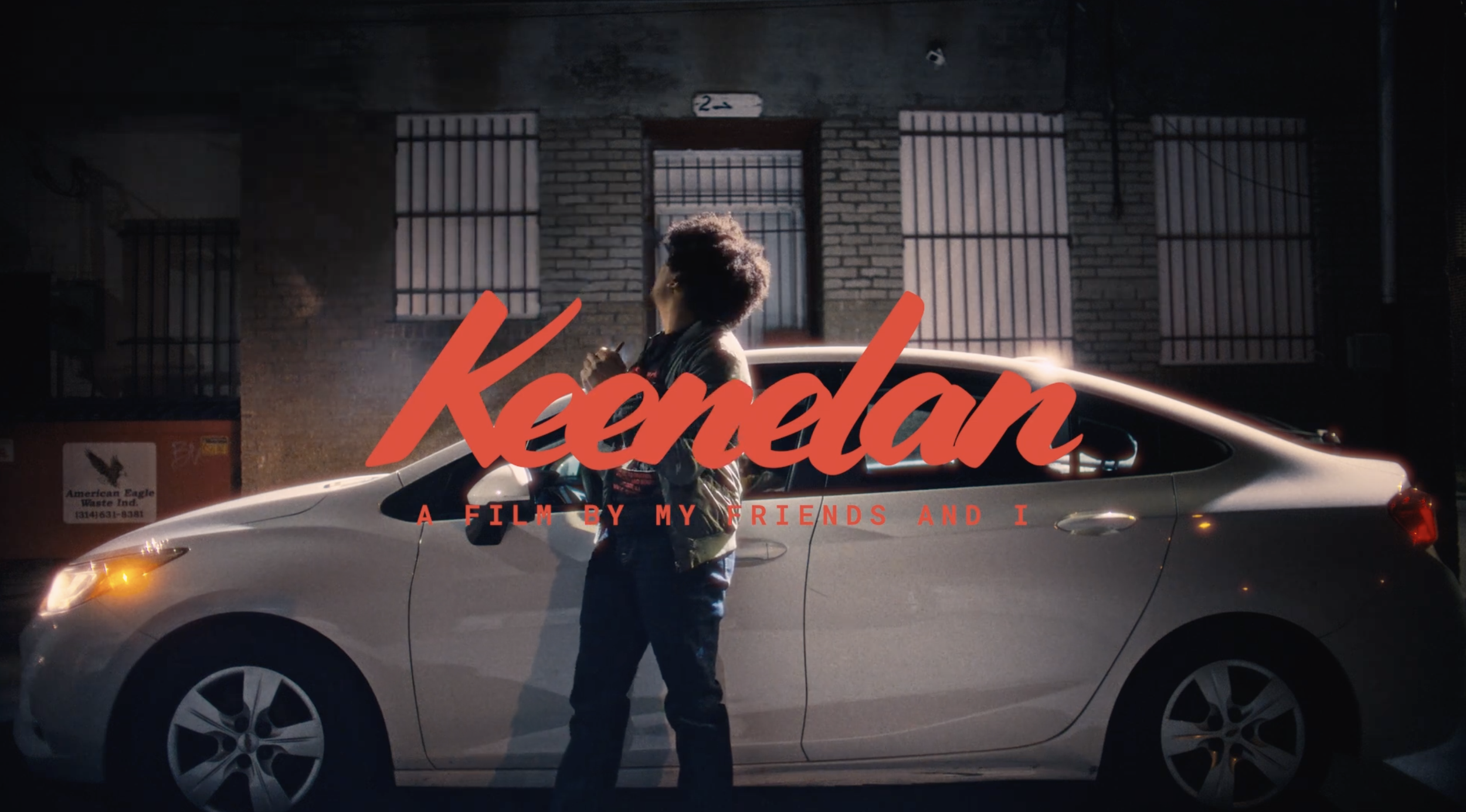 A person standing next to a silver car on a city street at night. The person has curly hair and is wearing a leather jacket, looking up. There is a brick building with barred windows behind them and a small sign with the number 2 above the doorway. The image features the title 'Kamden' in large red cursive font and the subtitle 'A film by my friends and I' in smaller red letters.