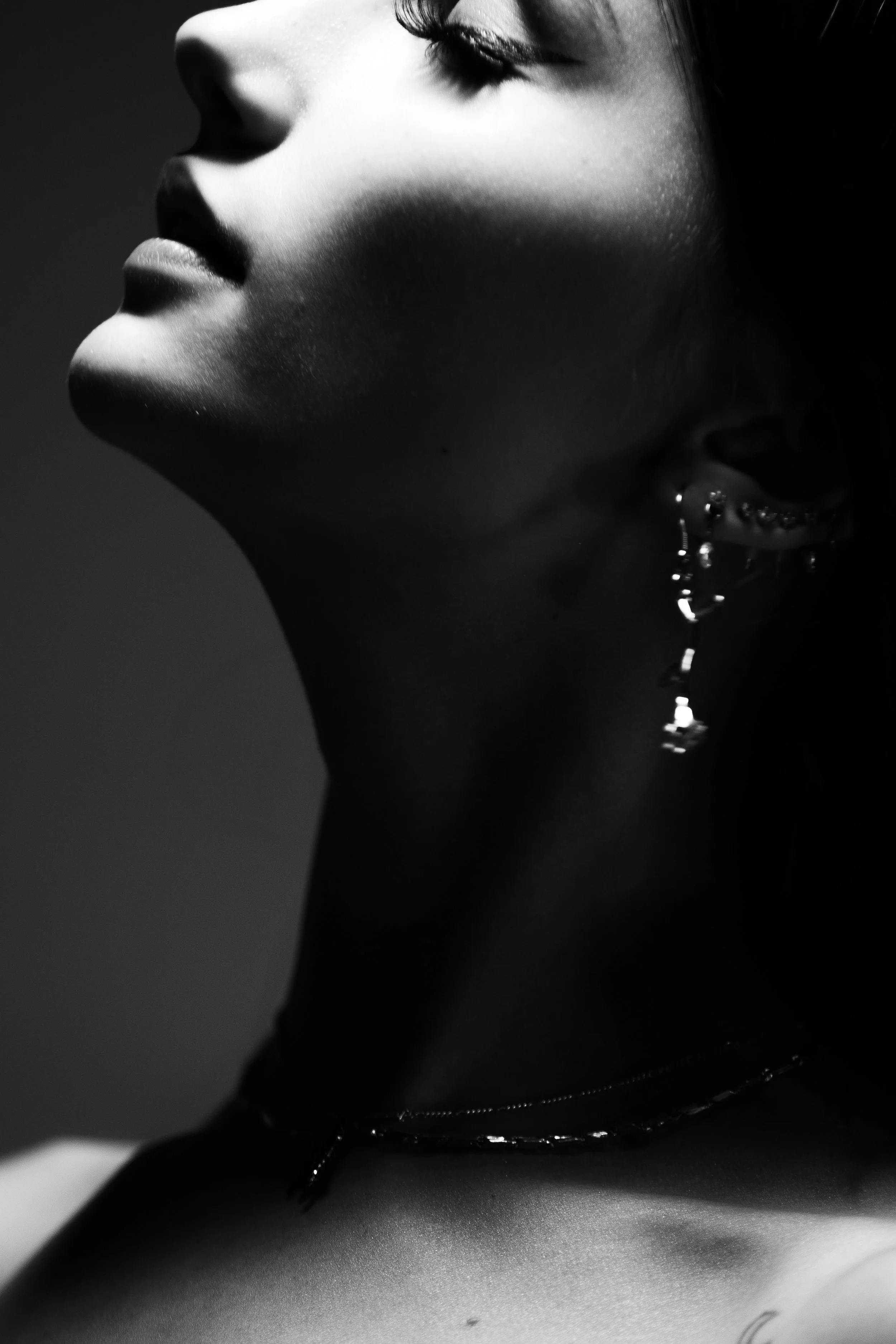 Close-up of a woman with her eyes closed, wearing earrings and a black necklace, with a shadowed background.