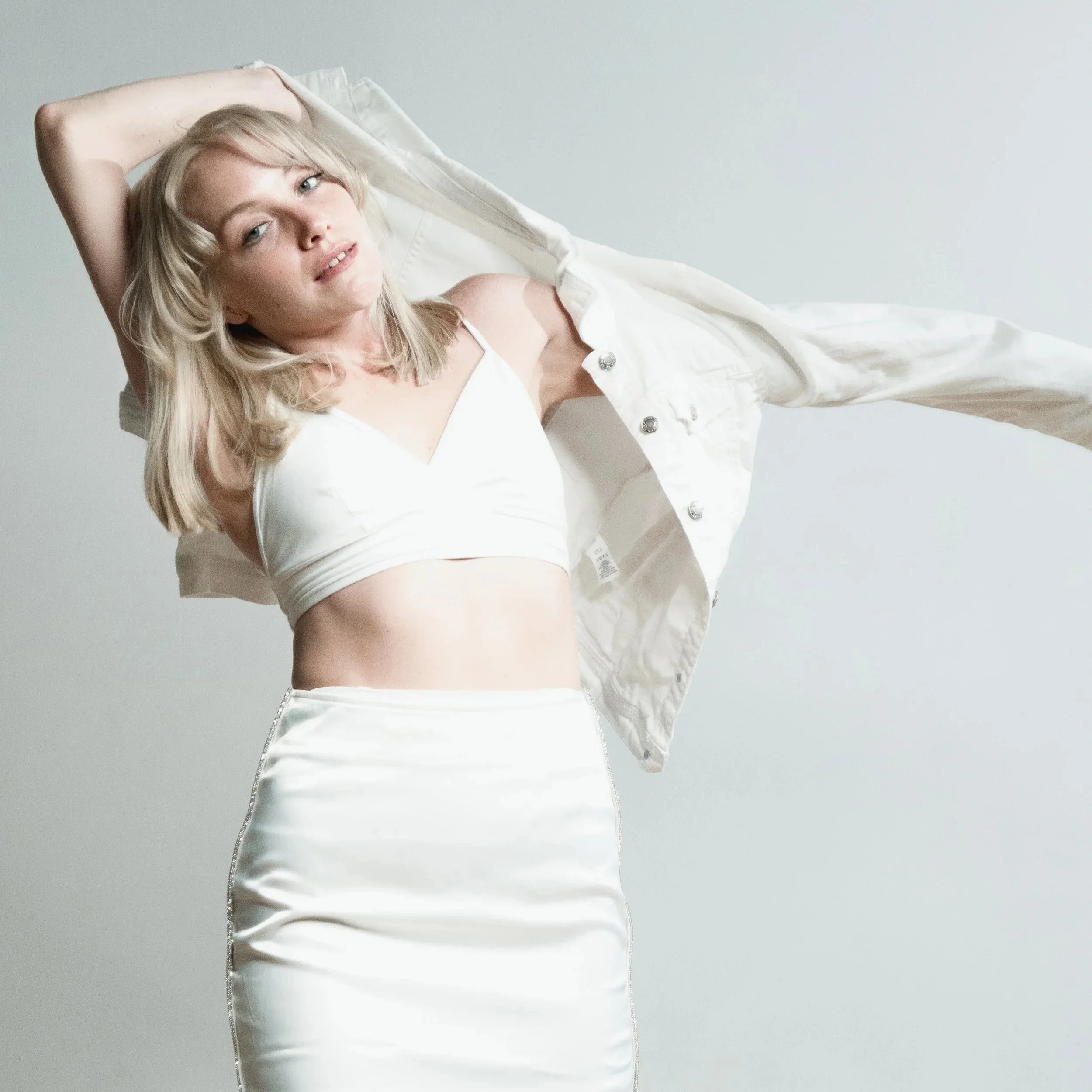 A young woman with blonde hair lying on her back with her arms above her head, wearing a white jacket and top, against a plain light gray background.