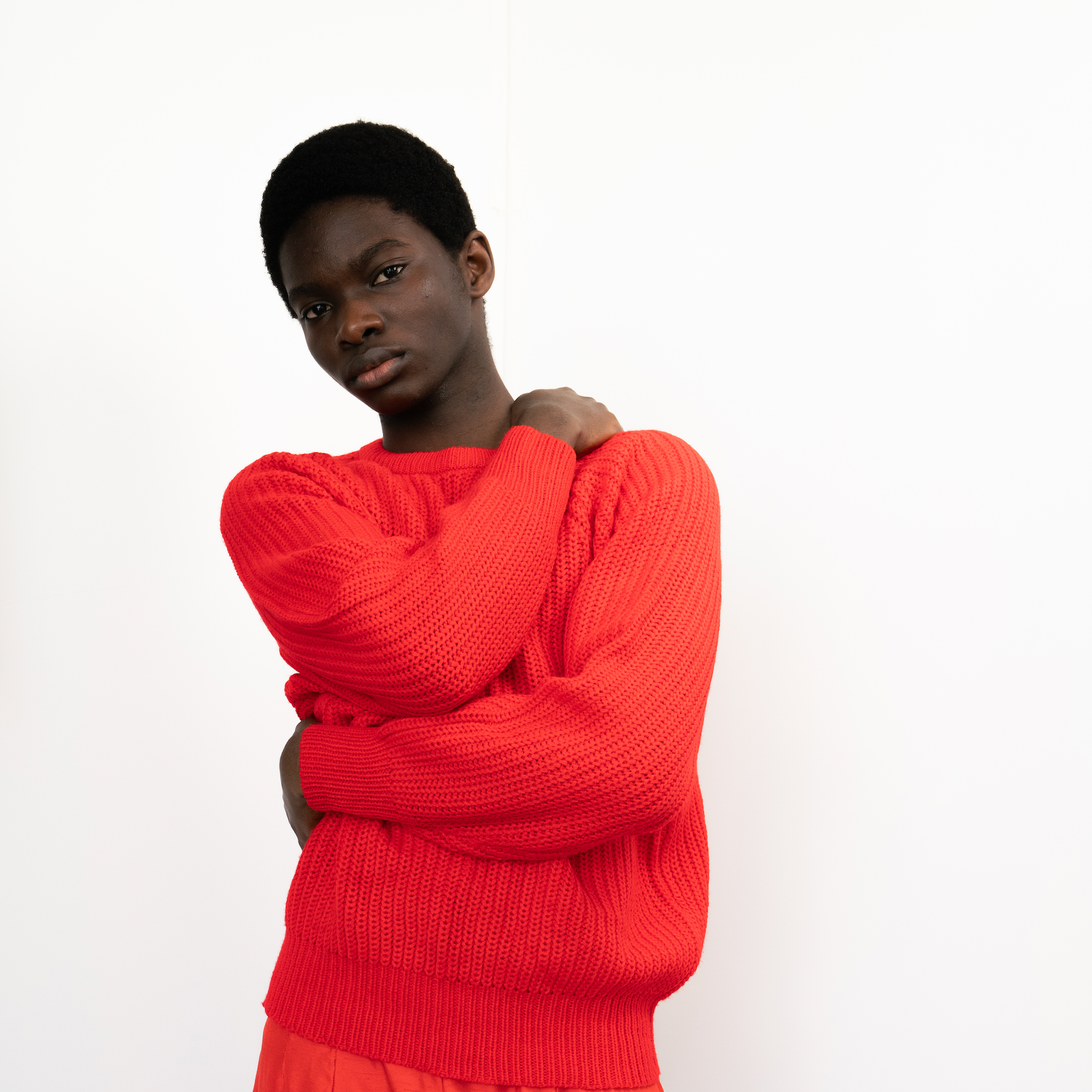 A young African American woman with short natural hair, wearing a bright red sweater, standing against a plain white background, with a serious expression and crossed arms.