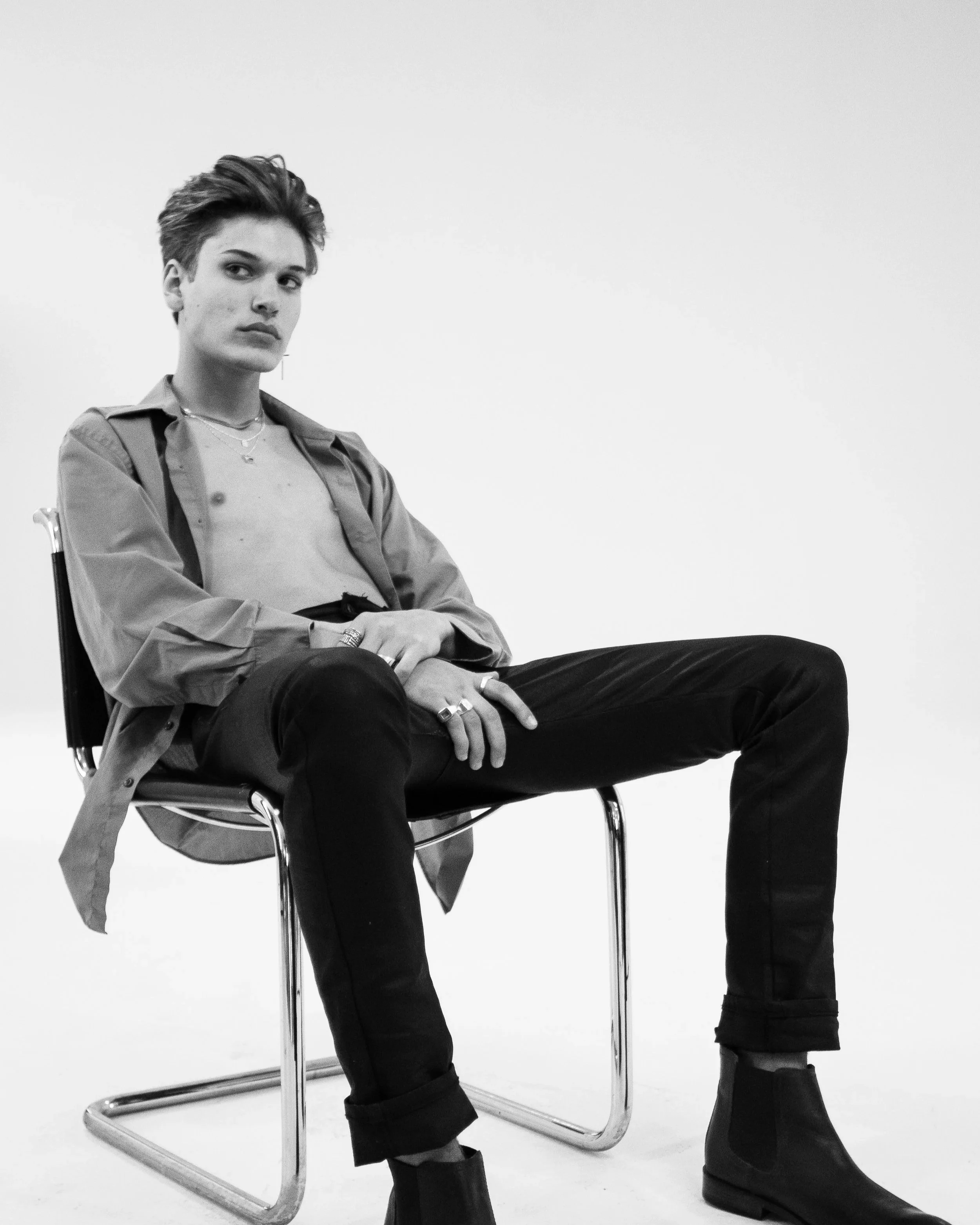 Black and white photograph of a woman with short hair sitting on a modern chair with a curved metal frame, wearing a jacket, t-shirt, and boots, against a plain white background.