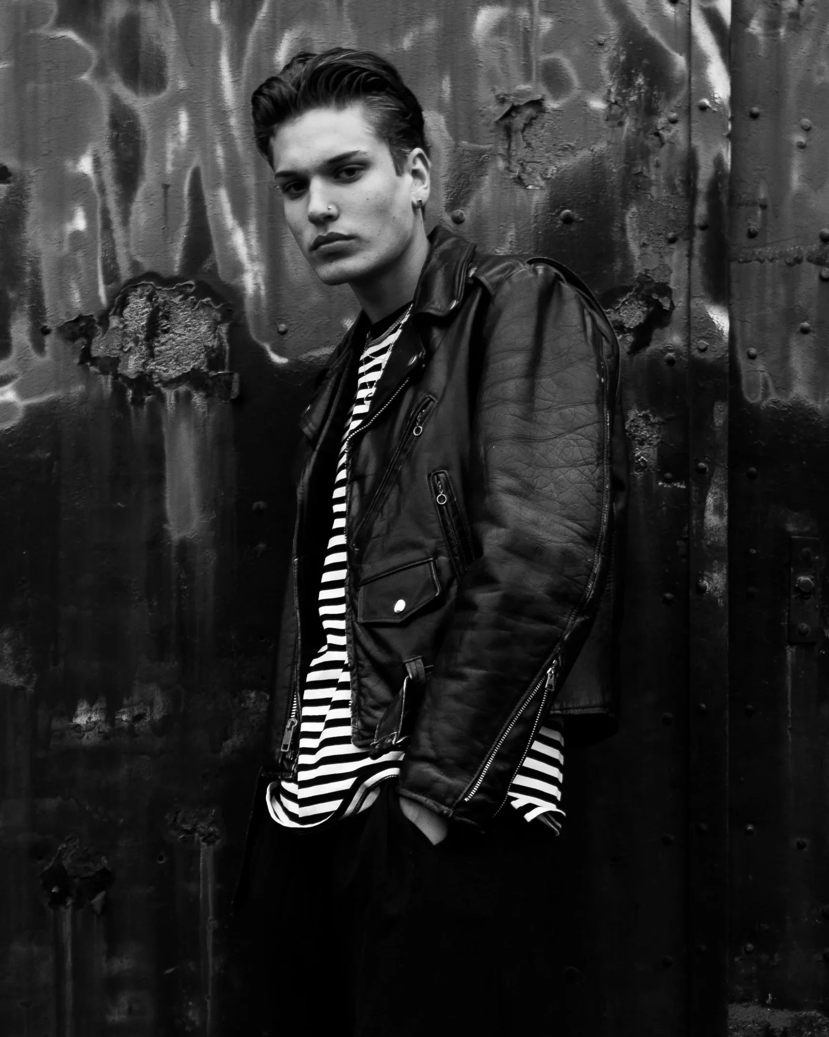A young person with short dark hair, wearing a leather jacket and striped shirt, standing in front of a textured metal wall.
