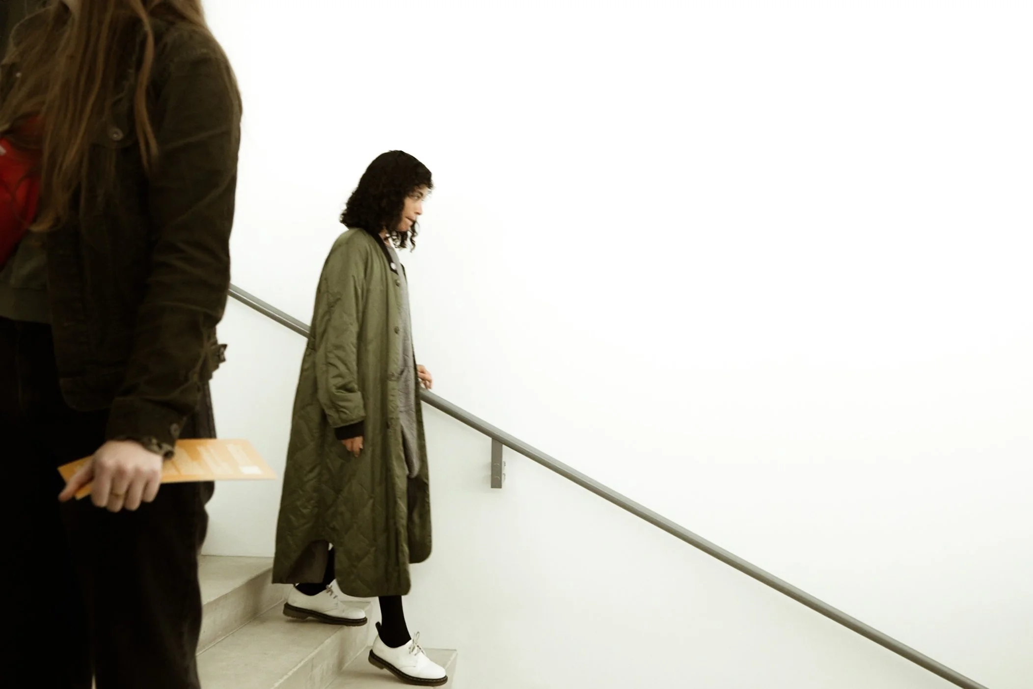 A woman in a beige coat and white shoes walking down stairs, holding the handrail, with another person partially visible in the foreground, in an indoor setting with plain white walls.
