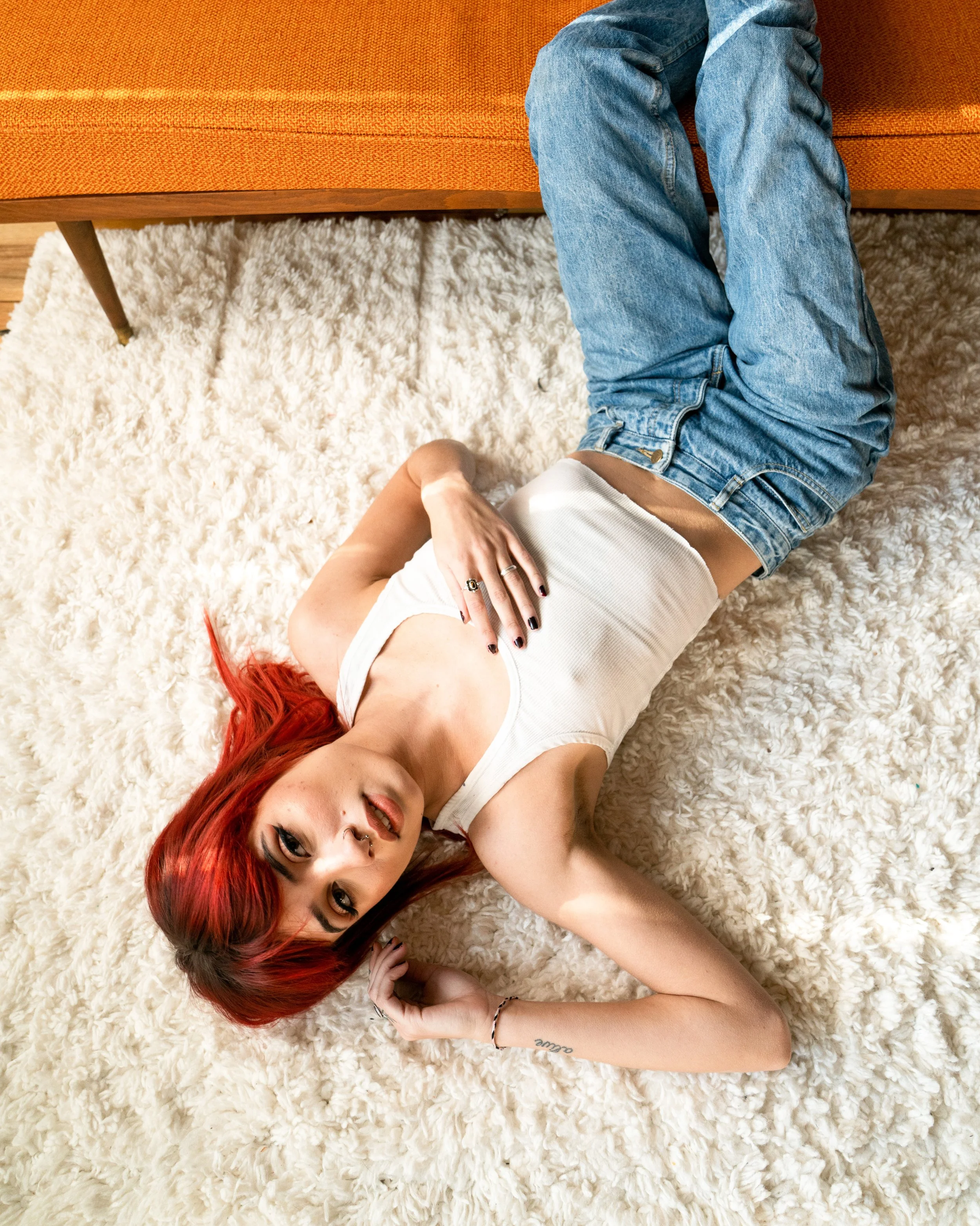 A woman with red hair lying on a fluffy white rug, wearing a white tank top and jeans, with her hand resting on her stomach, and looking at the camera.