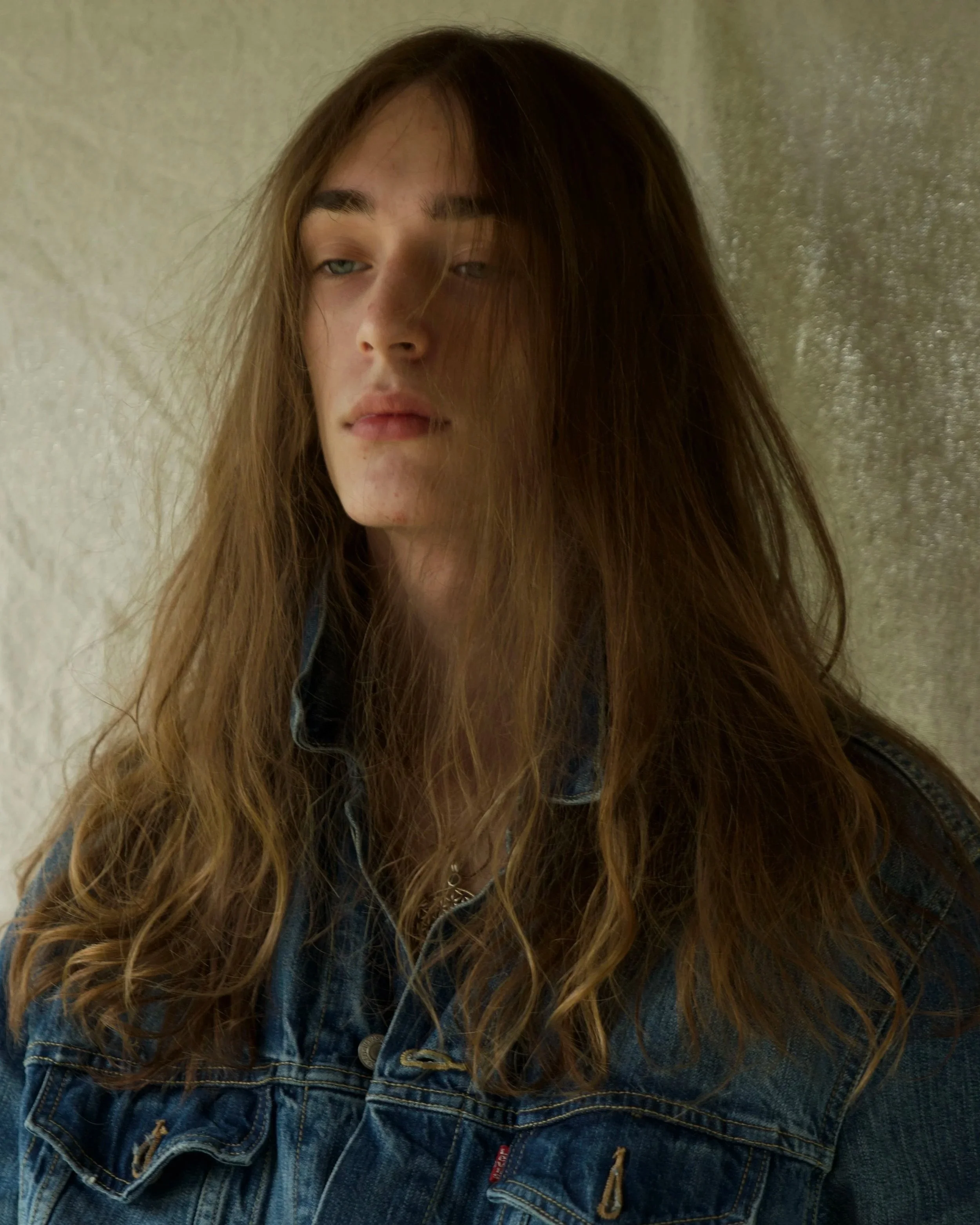Close-up of a young woman with long, wavy brown hair wearing a denim jacket with a small red tag, standing against a textured beige wall.