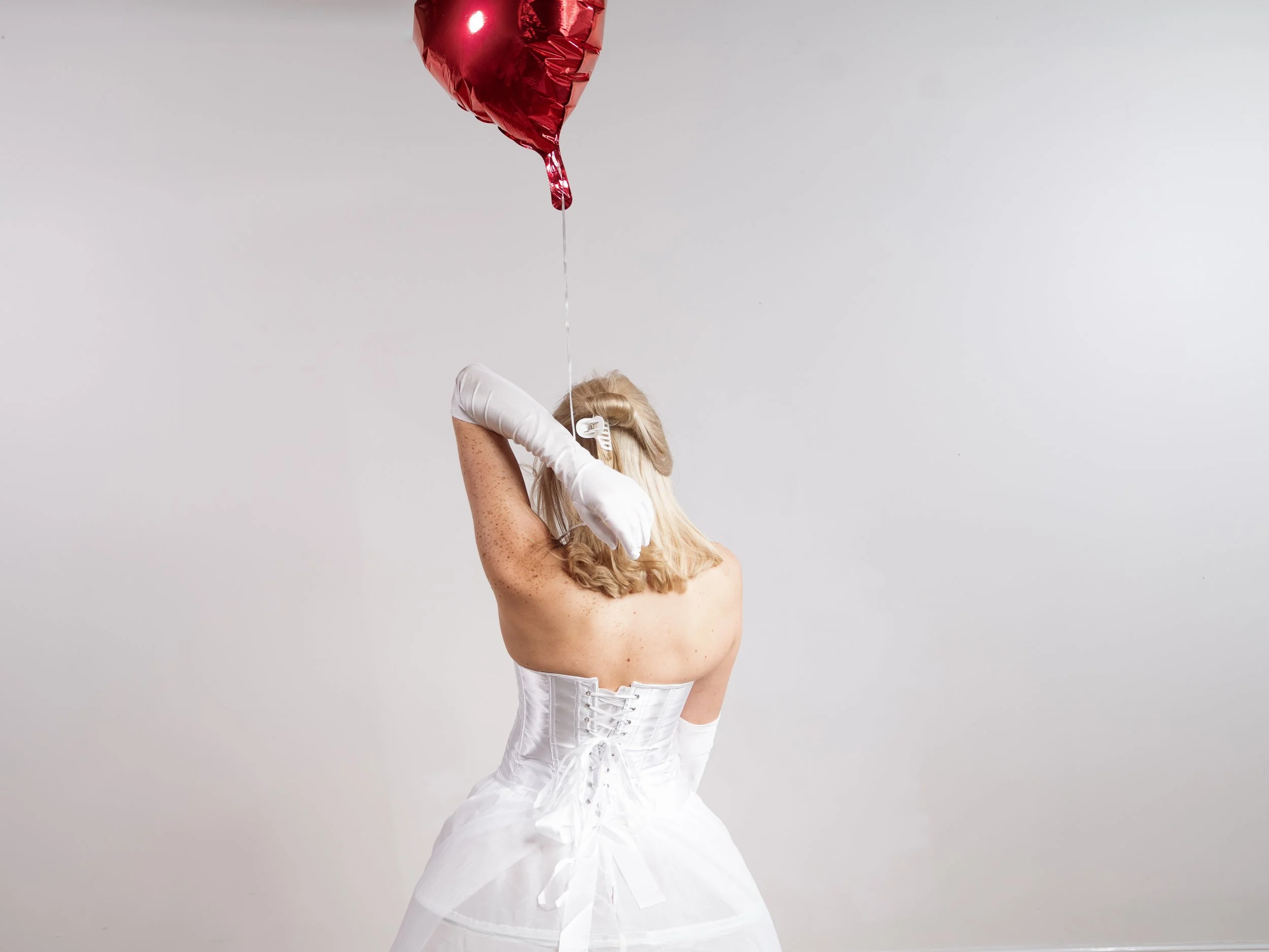 A woman with long blonde hair wearing a white corset and white gloves, holding a red heart-shaped balloon