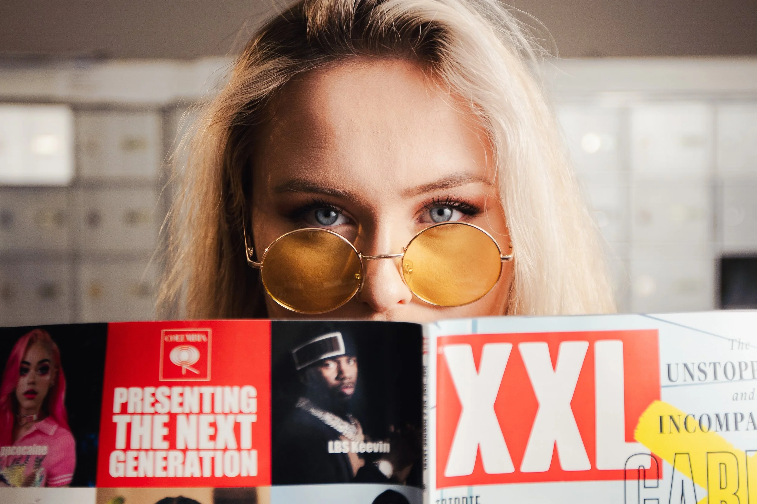 A person with blonde hair and blue eyes wearing round yellow-tinted sunglasses is looking over a magazine, which is partially covering their face. The magazine cover features a woman with bright pink hair and another with a serious expression.