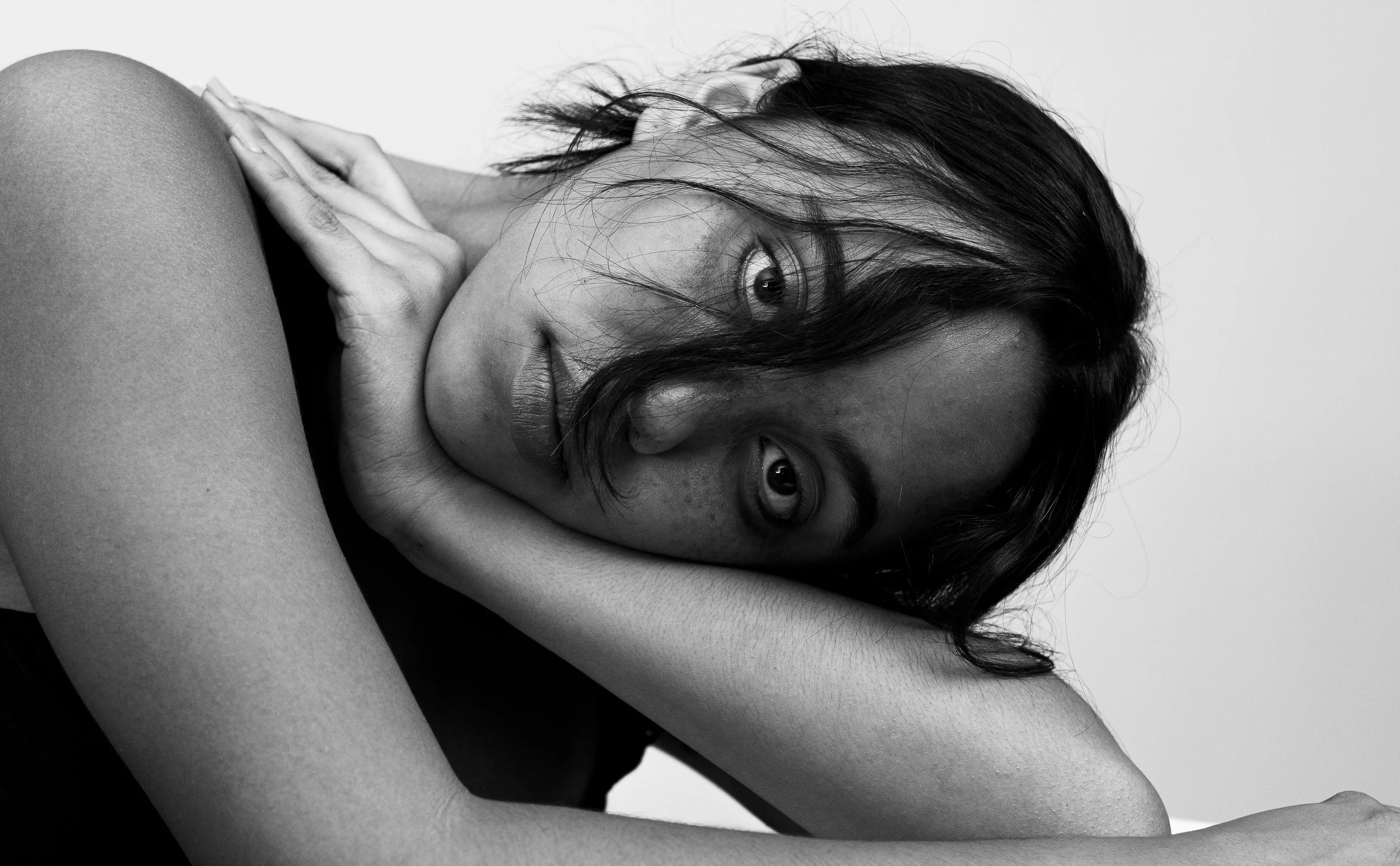 A black and white photograph of a woman with dark, slightly messy hair. She is resting her head on her forearm, looking directly at the camera with a contemplative expression. Her skin is smooth with a few visible freckles, and her eyes are wide and 