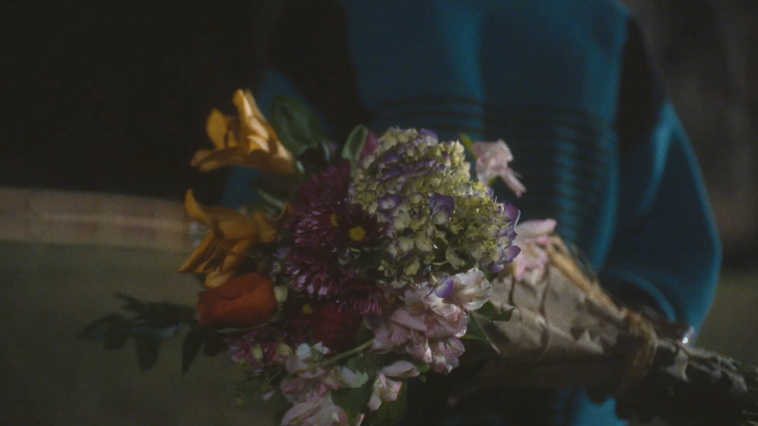 A person wearing a blue jacket holding a bouquet of colorful flowers with purple, yellow, and pink blooms, in a dimly lit setting.
