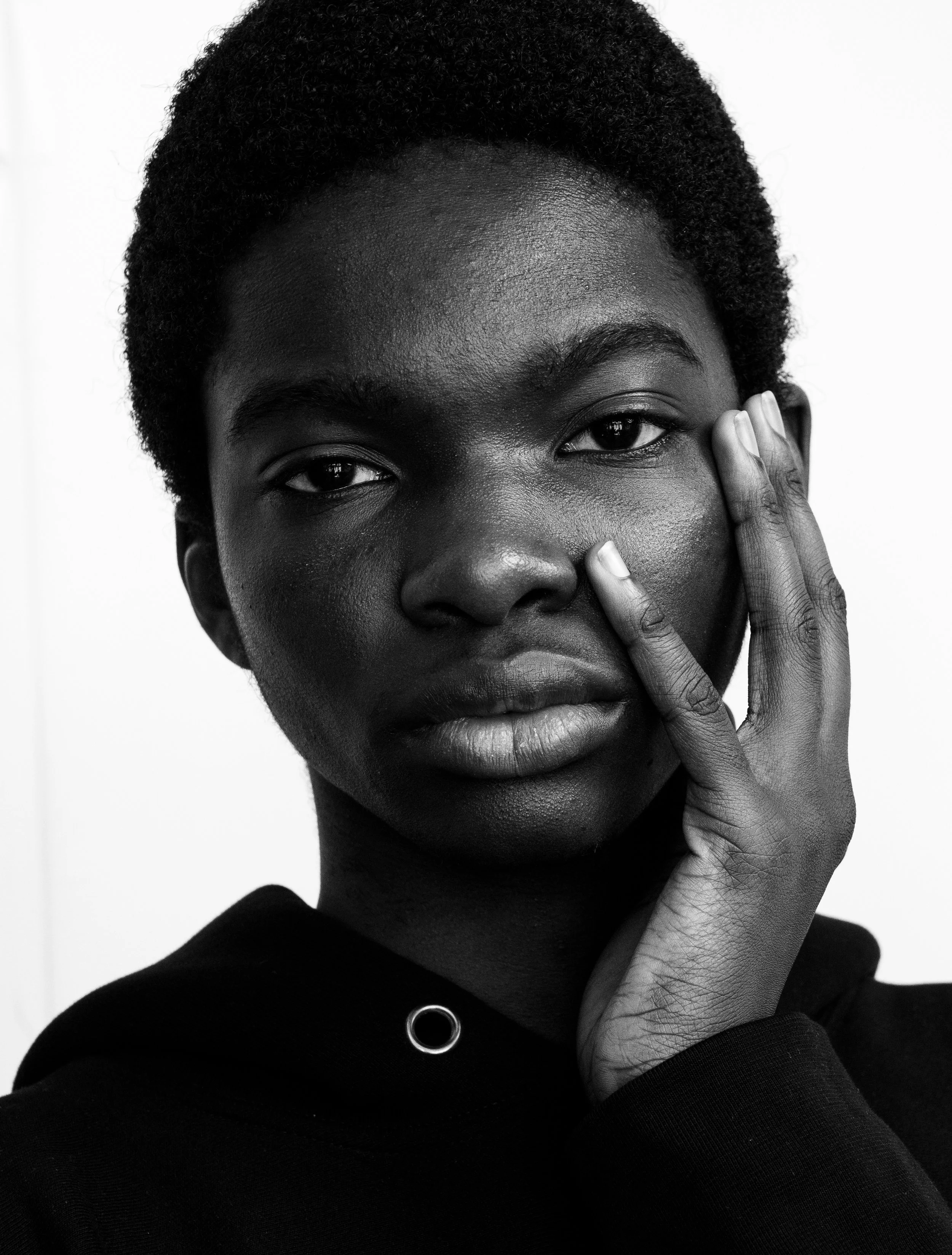 A black and white photograph of a person with dark skin and short, curly hair. They are touching their face with their right hand, with fingers near their cheek and thumb near their chin. The person is wearing a dark top with a visible circular detai