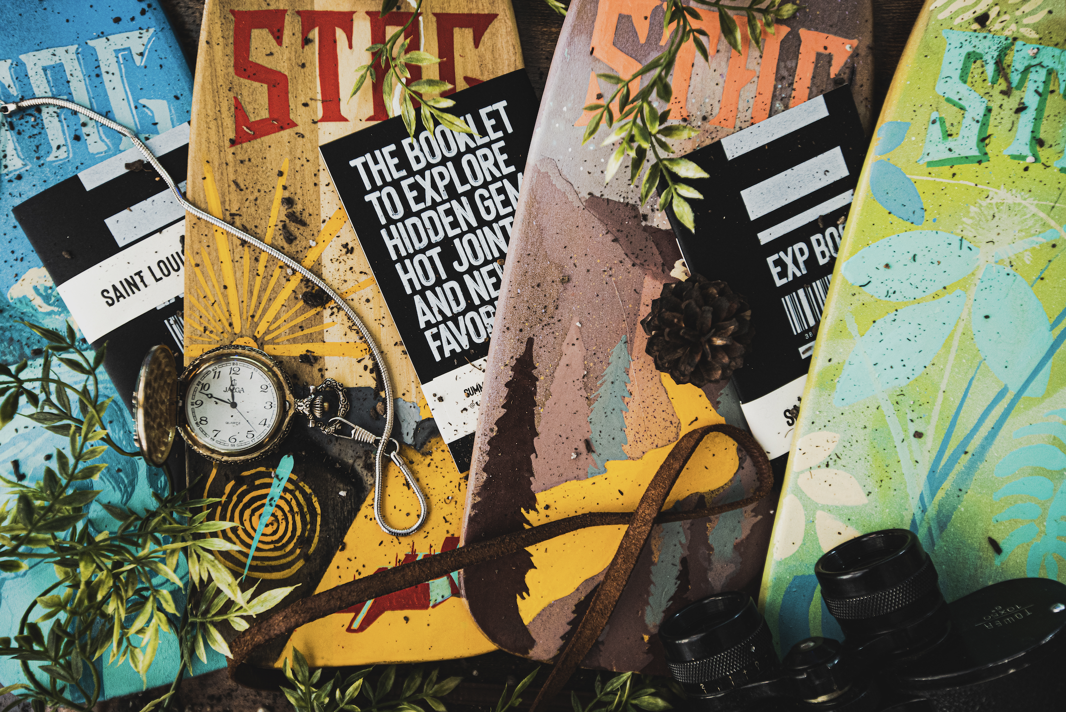 A collection of colorful guidebooks, a pocket watch, a necklace, leaves, a pine cone, and a small telescope on a dark surface.