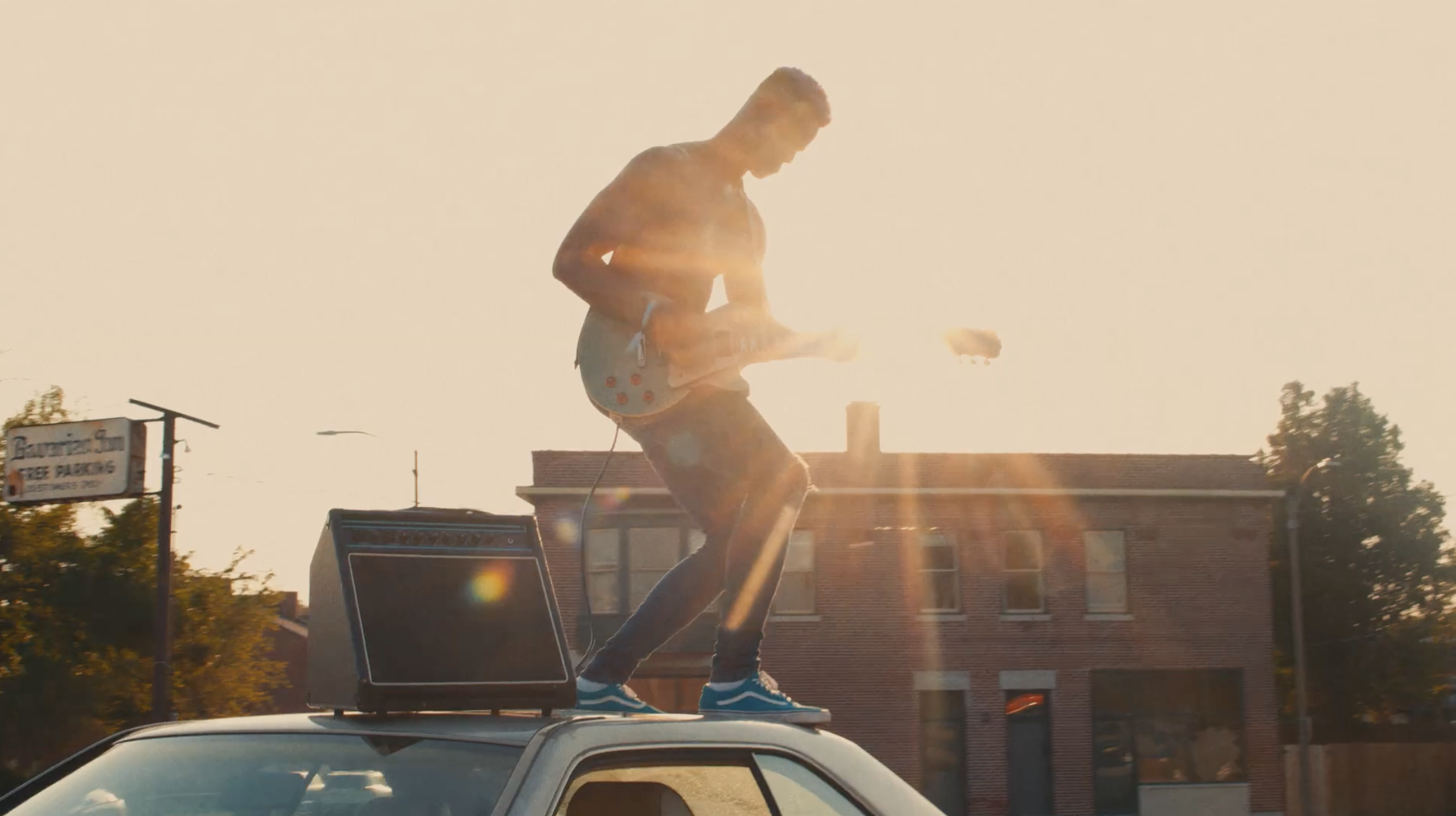 A young man playing an electric guitar on top of a car during sunset, with buildings and trees in the background.