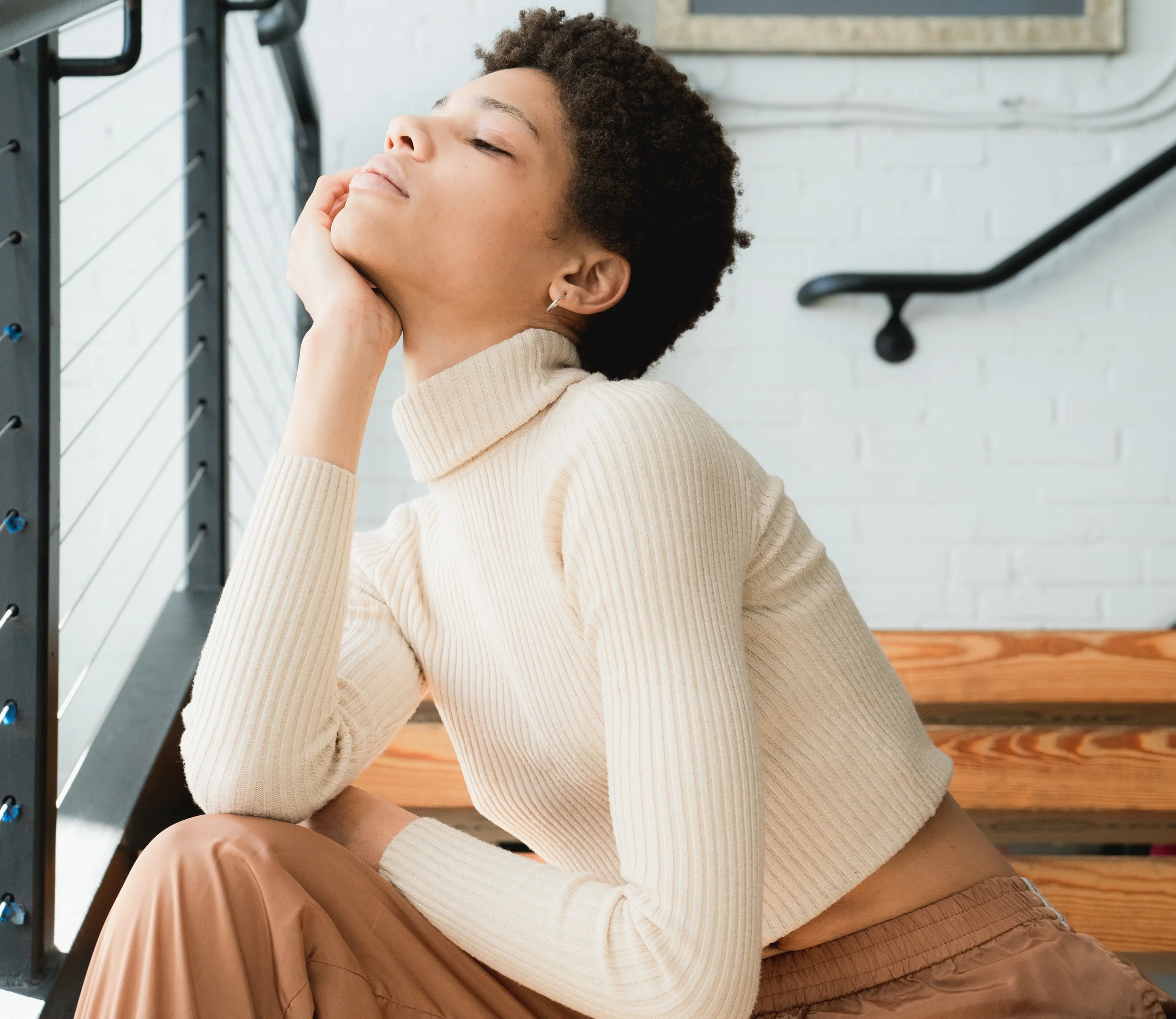 A person with short curly hair and earrings wearing a white ribbed turtleneck sweater, sitting against a background with a white brick wall, a black metal shelf, and wooden flooring.