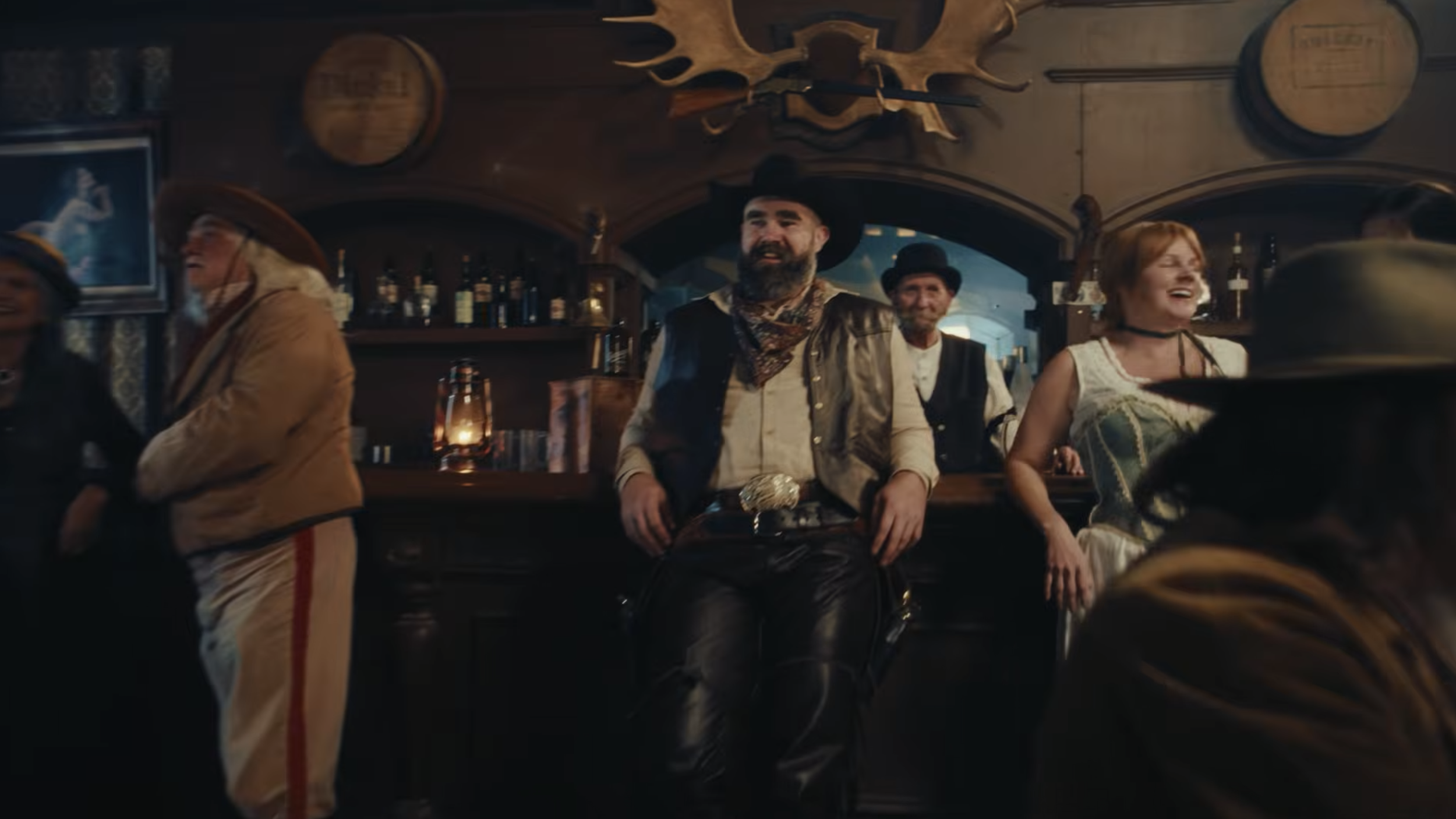 Group of people dressed in old Western costumes in a rustic bar, with a man with a beard and cowboy hat sitting at the counter, laughing, and others standing and smiling around him.