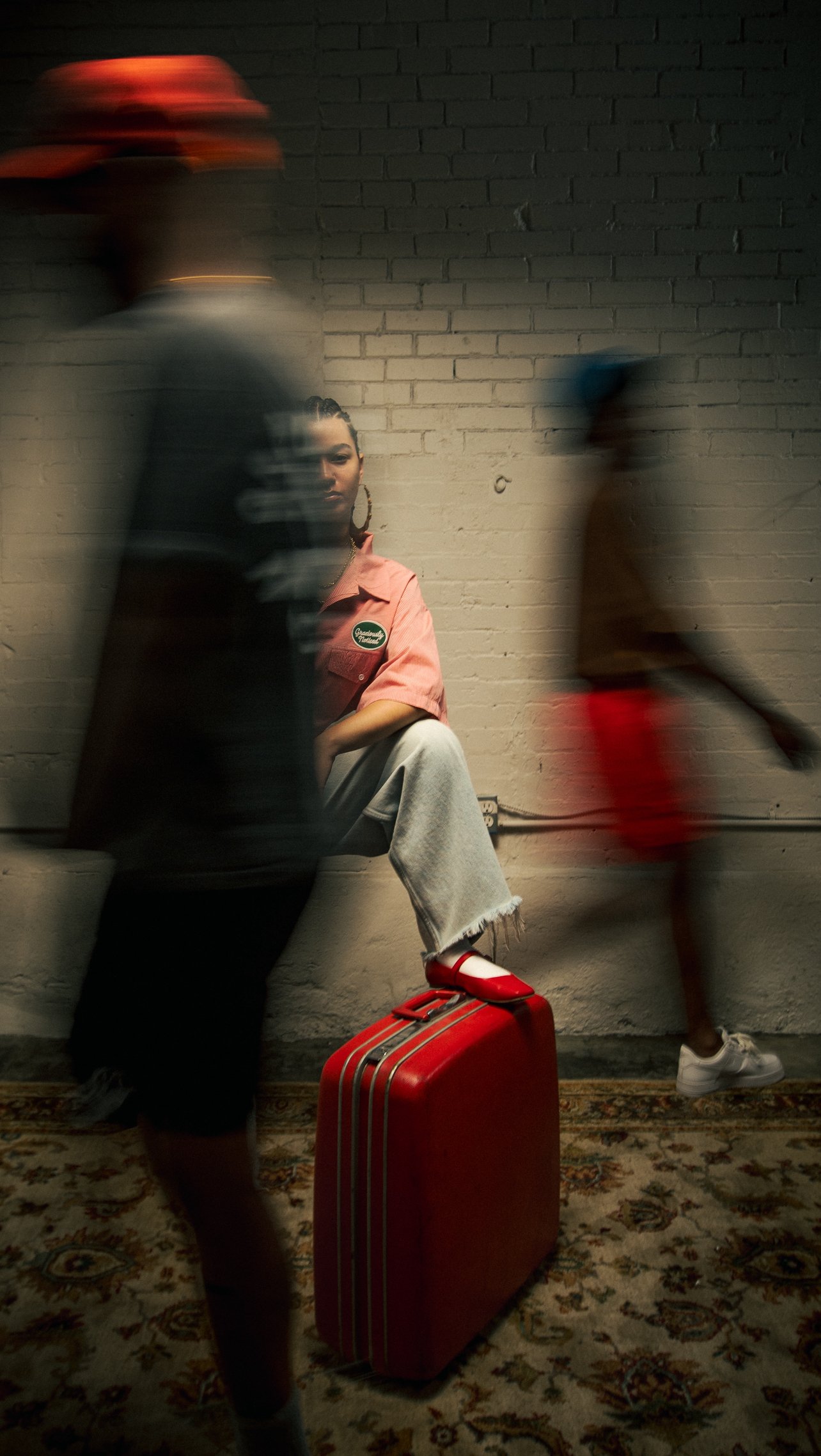 A woman sitting on a bench with one leg crossed over the other, holding a book and a pink shirt with an emblem. She has large hoop earrings and curly hair. A red suitcase is on the floor beside her. In motion, a person with a red hat and a person in 