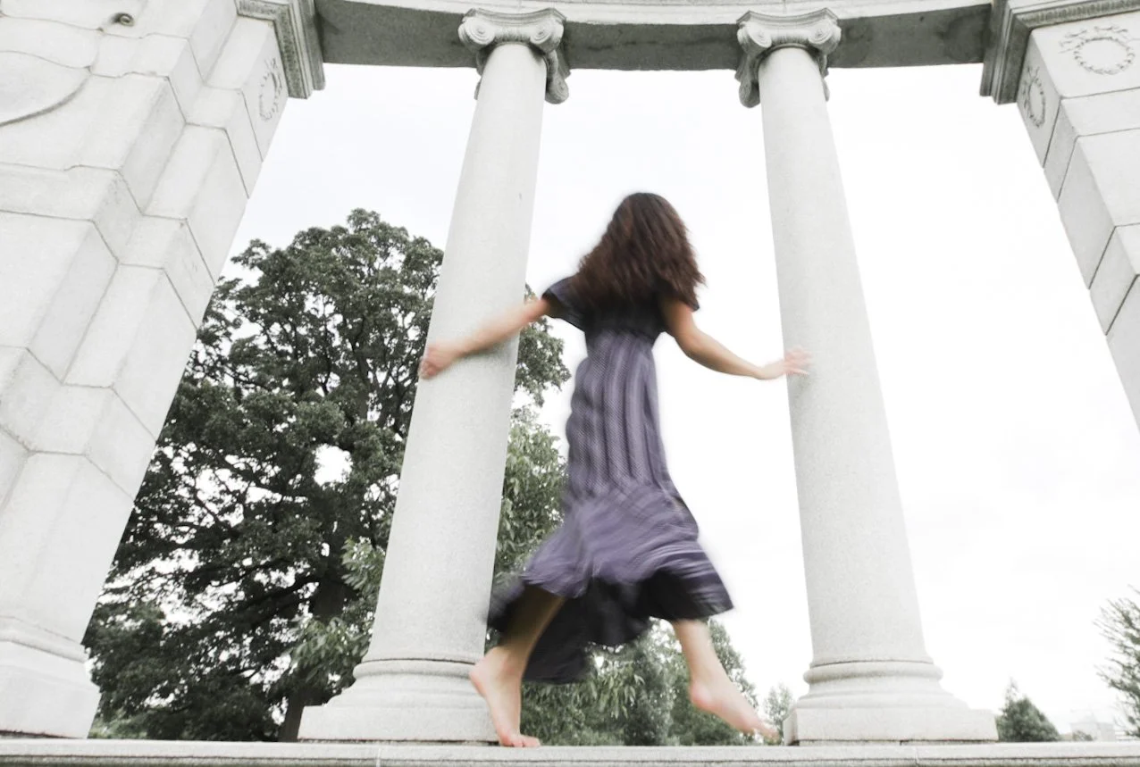 A woman in a long dress is walking on a stone platform with large stone columns and an arch above, with trees in the background.