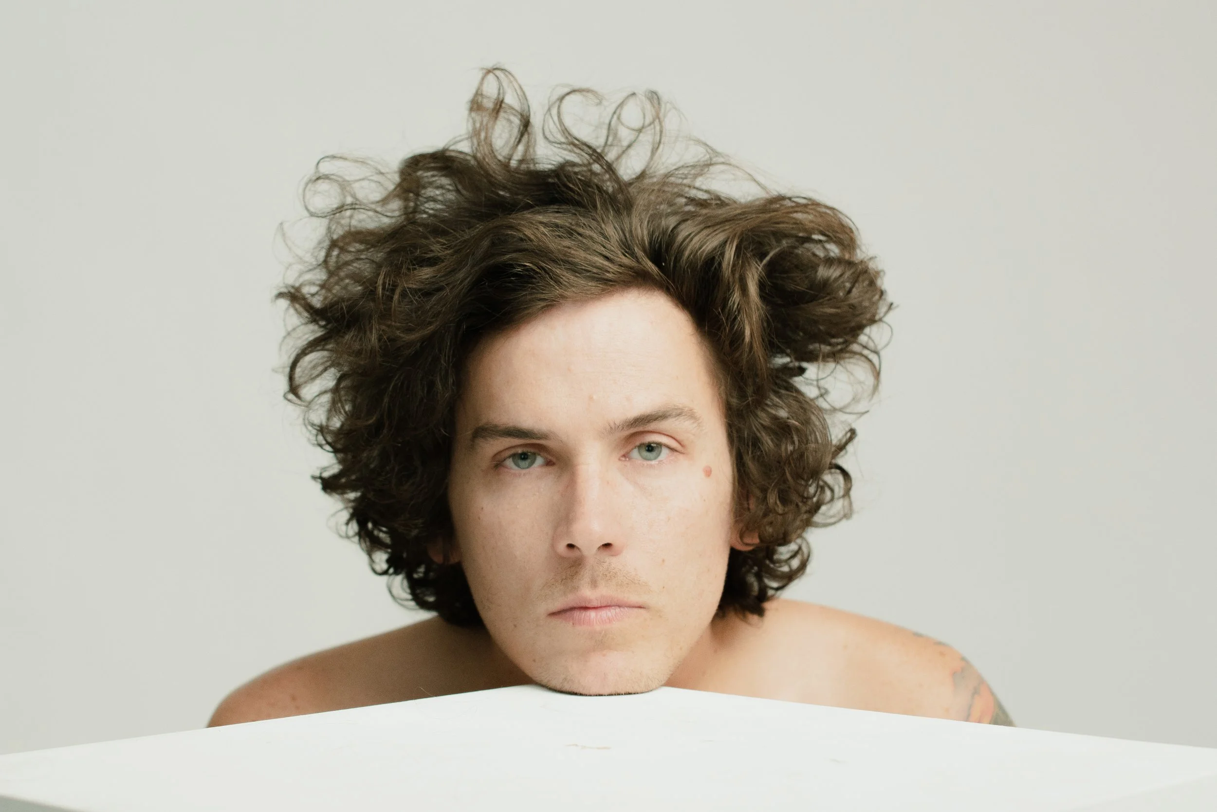 A person with curly brown hair and piercing blue eyes looking directly at the camera, with a plain light background.