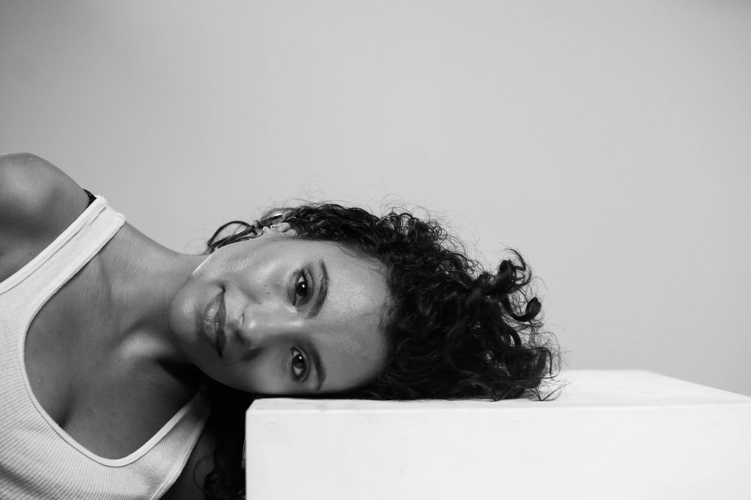 A black and white photo of a woman with curly hair resting her head on a white block, wearing a sleeveless top and looking at the camera.