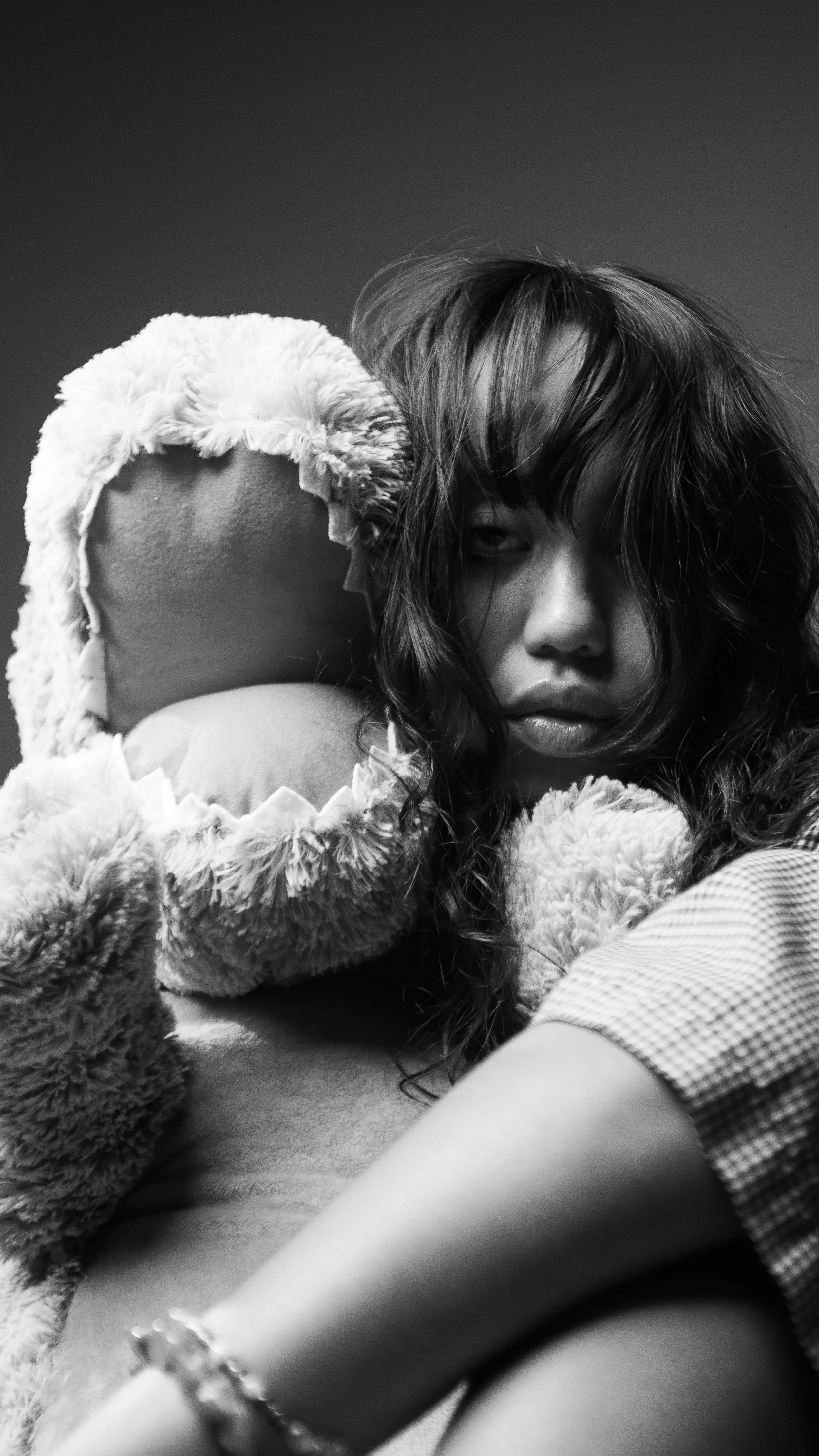 A young woman with tousled hair and a serious expression, holding a fluffy teddy bear, in a black and white close-up portrait.