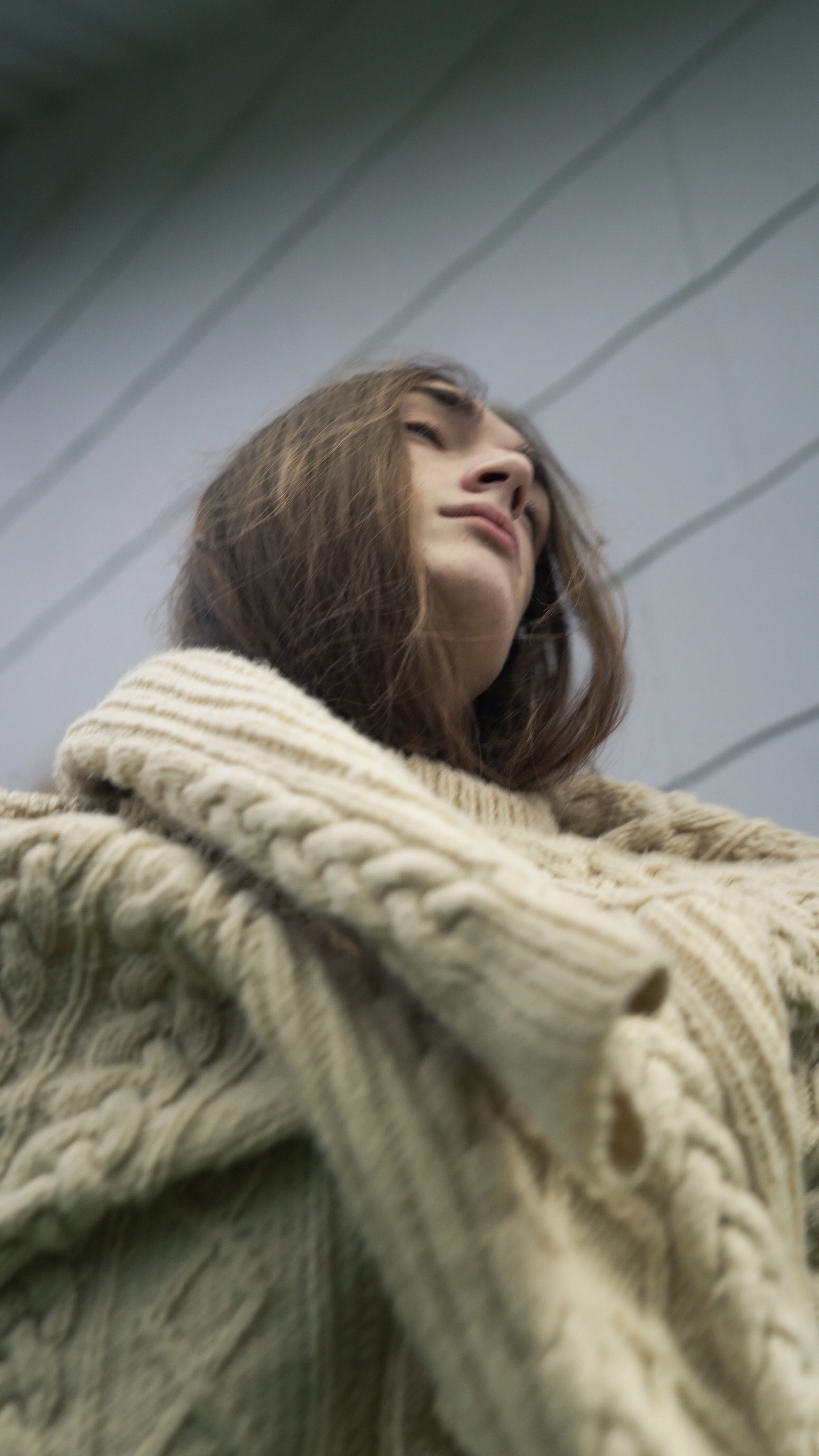 Close-up of a young woman with shoulder-length brown hair, wearing a beige knitted sweater, looking downward with a neutral expression, against a background of ceiling panels.