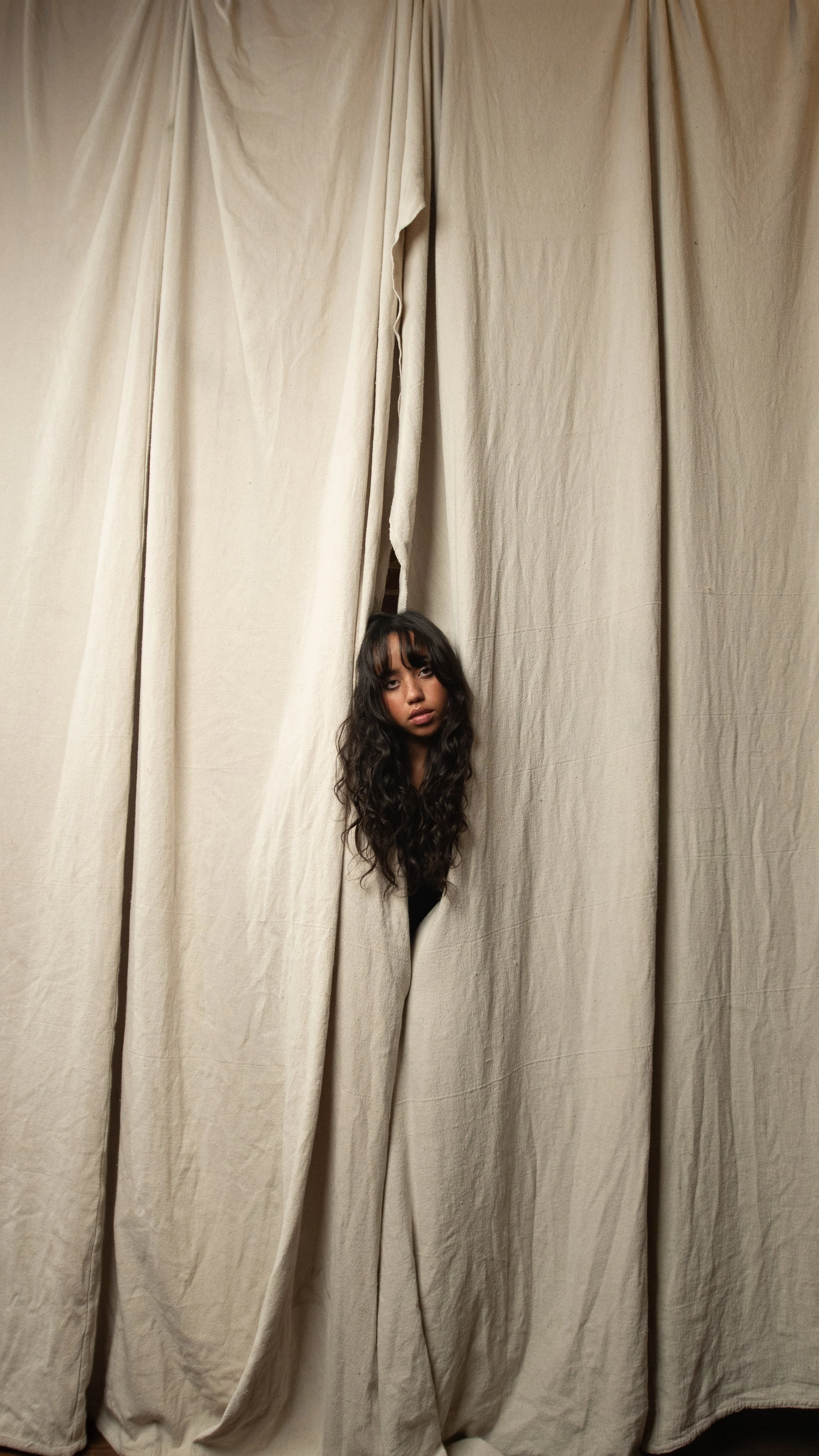 A young woman with long, curly dark hair and bangs peeking through a gap in beige curtains.