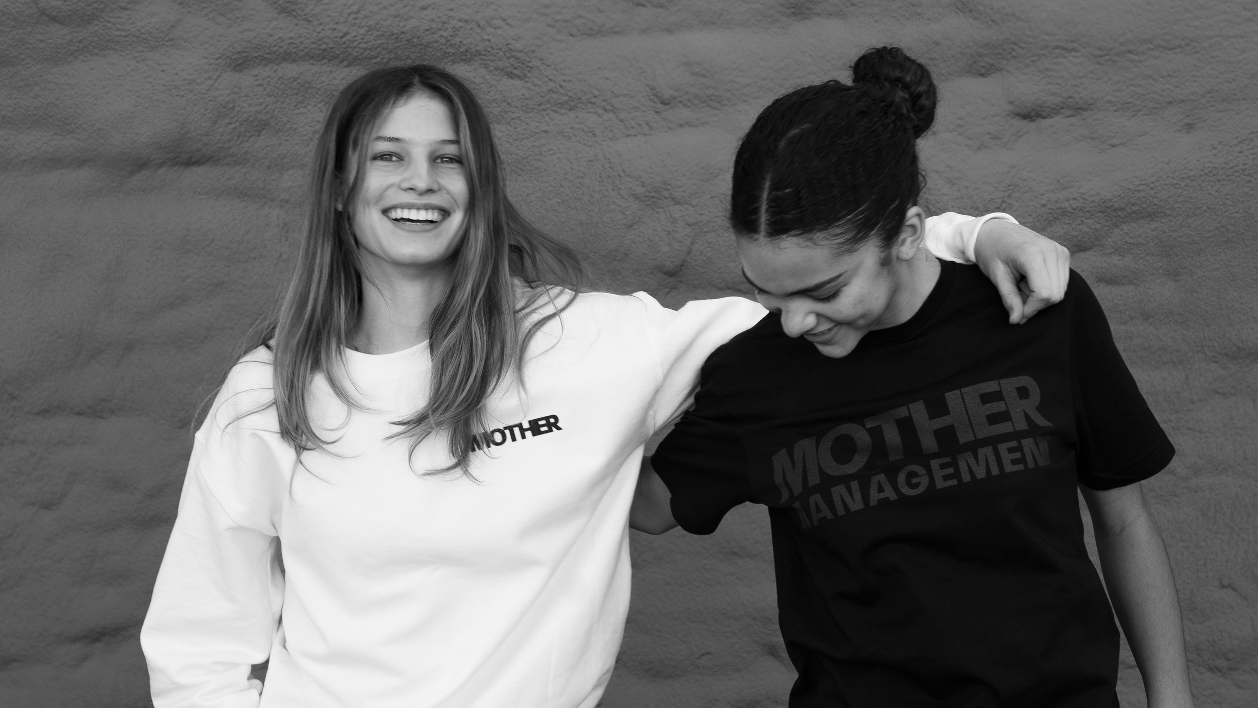 Two young women are standing together against a textured wall, with one smiling at the camera and the other looking down. They have their arms around each other, indicating a close relationship.