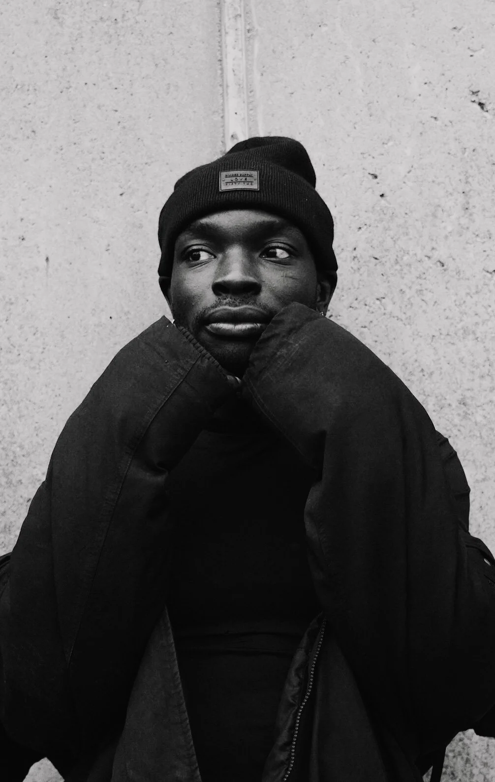 A black and white photo of a man wearing a beanie and a jacket, sitting against a textured wall. He has a thoughtful expression and is resting his chin on his hands.