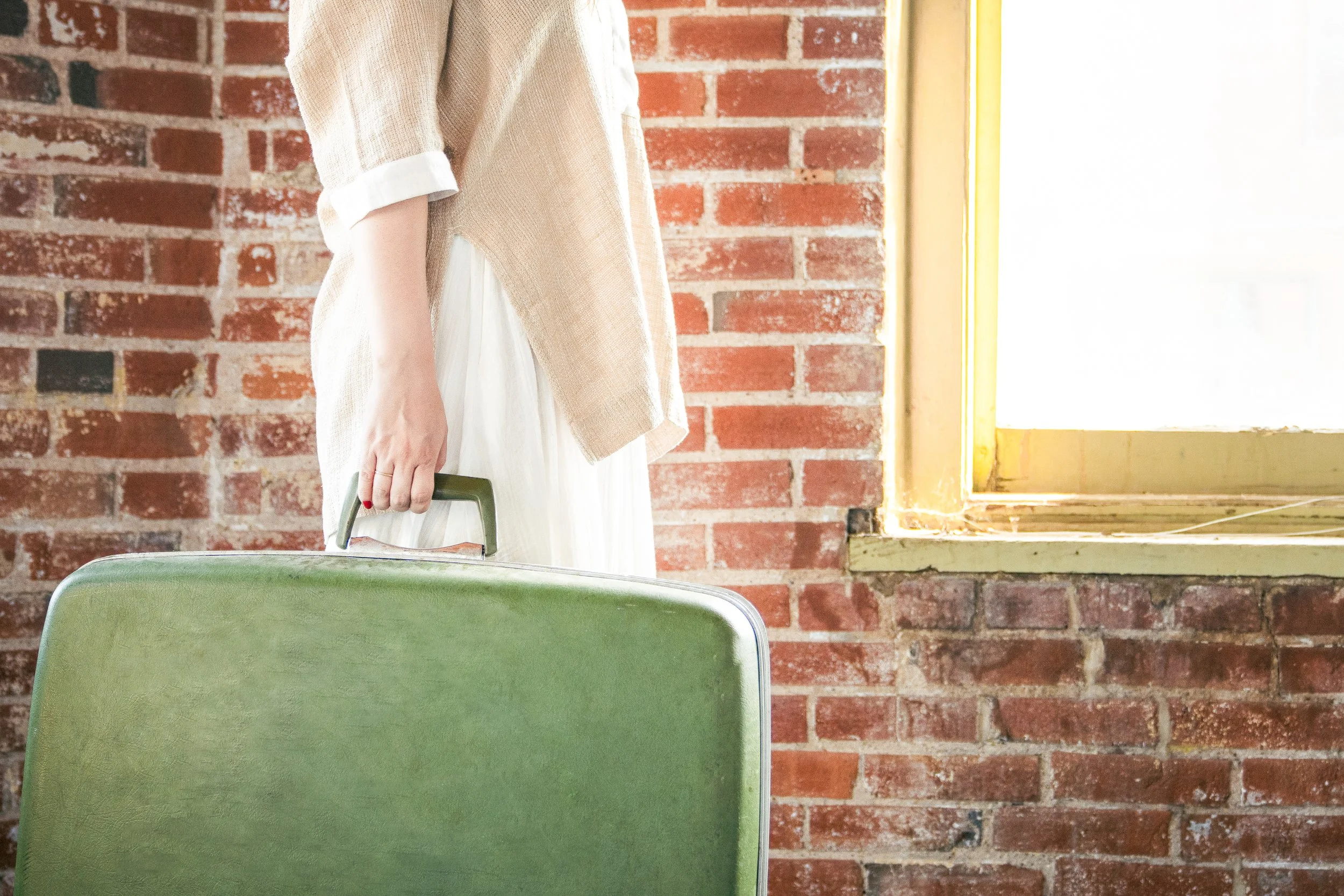 Part of a person wearing a beige coat and white dress standing next to a green chair, holding a small suitcase, in front of a red brick wall and a yellow-framed window.
