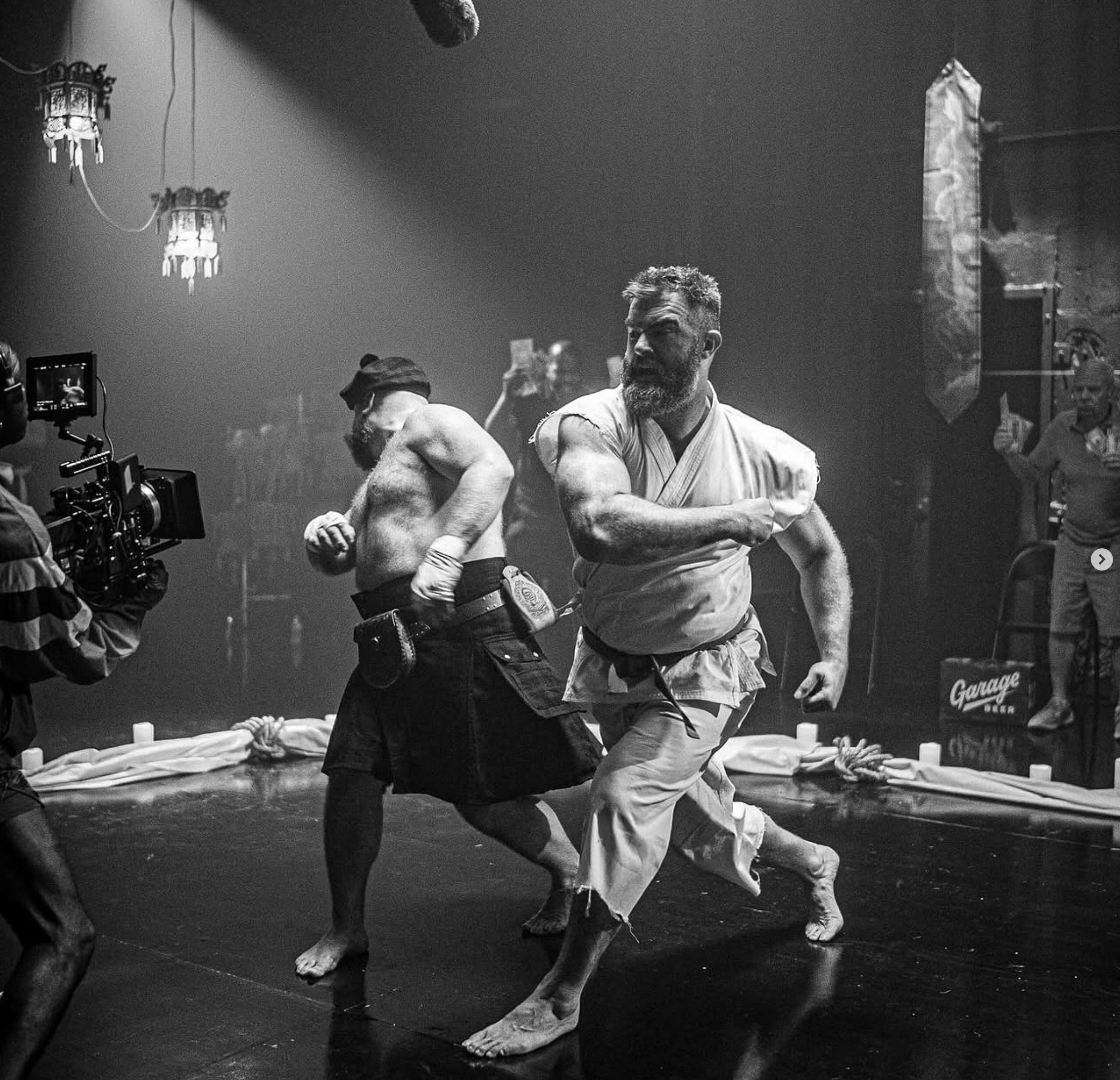 Two men practicing martial arts, wearing traditional white uniforms, are standing on a mat in a dojo or training room. One man is looking to his right, while the other is facing away. There are lights hanging from the ceiling and other people in the 