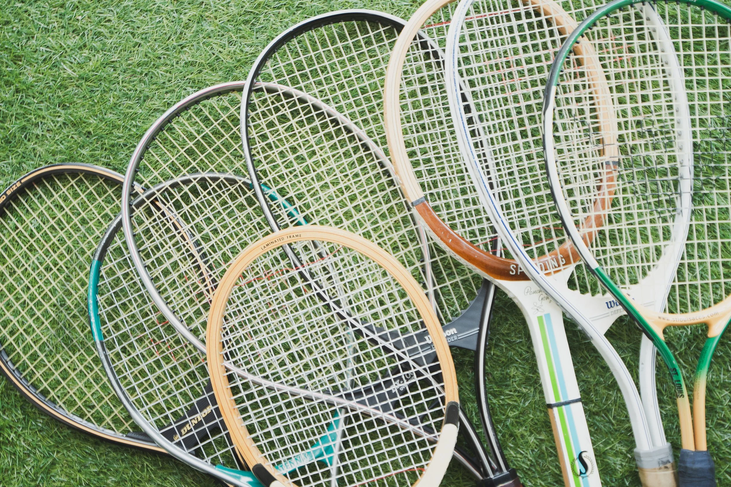 Multiple tennis rackets of various colors and brands lying on green grass.