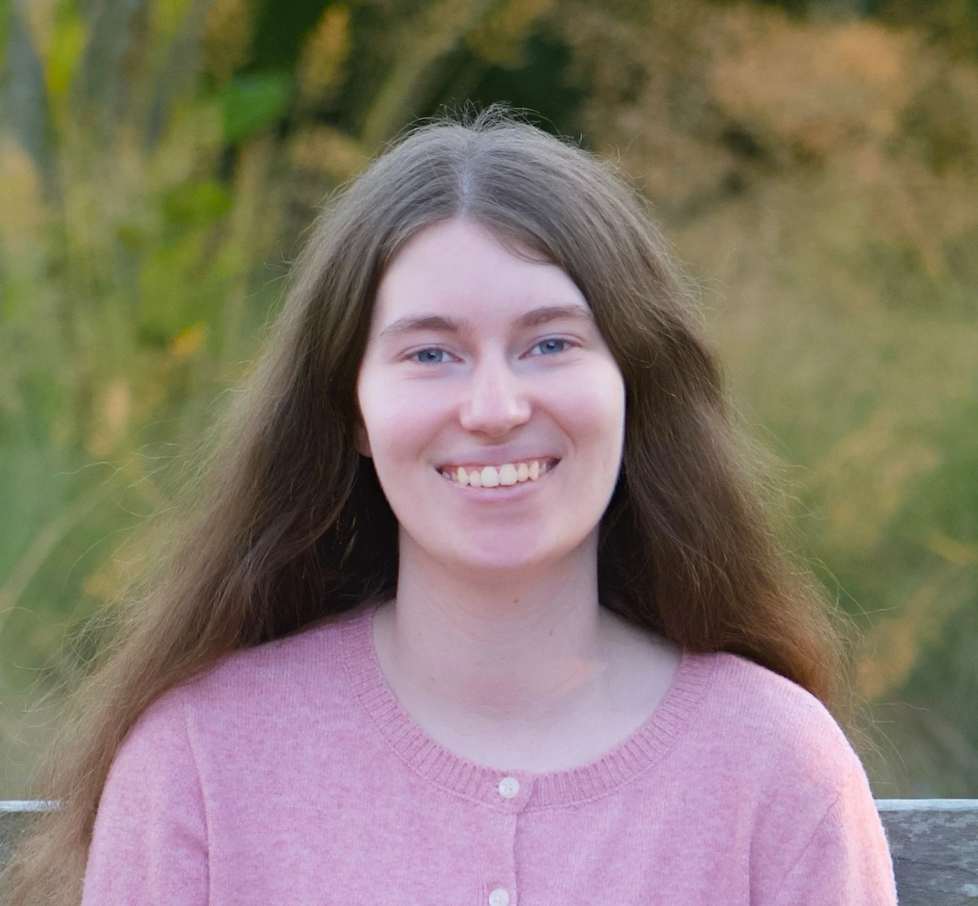 Smiling woman with long brown hair wearing a pink sweater outdoors