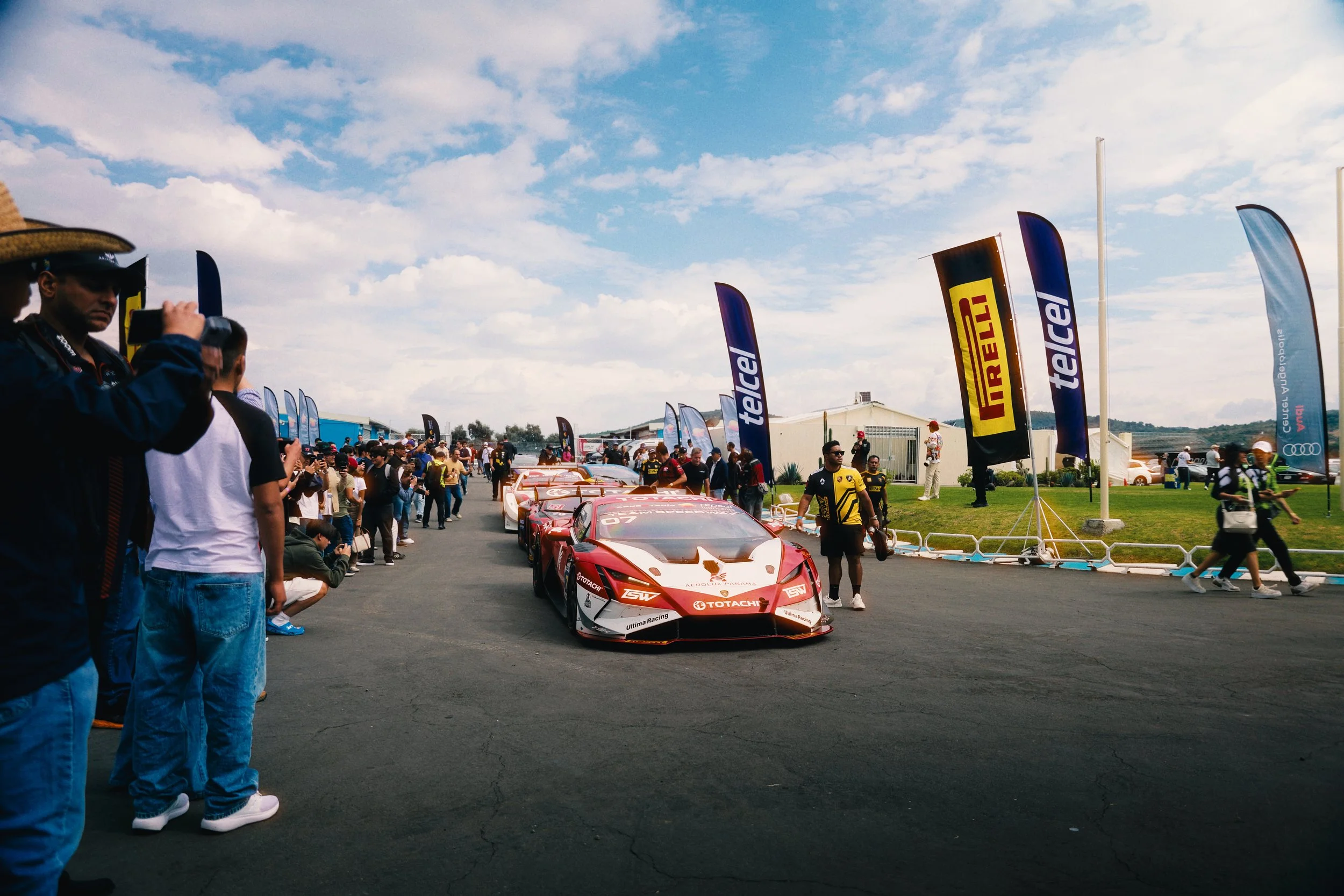Race cars on display at an outdoor event with a crowd of spectators and flags from various sponsors, under a partly cloudy sky.