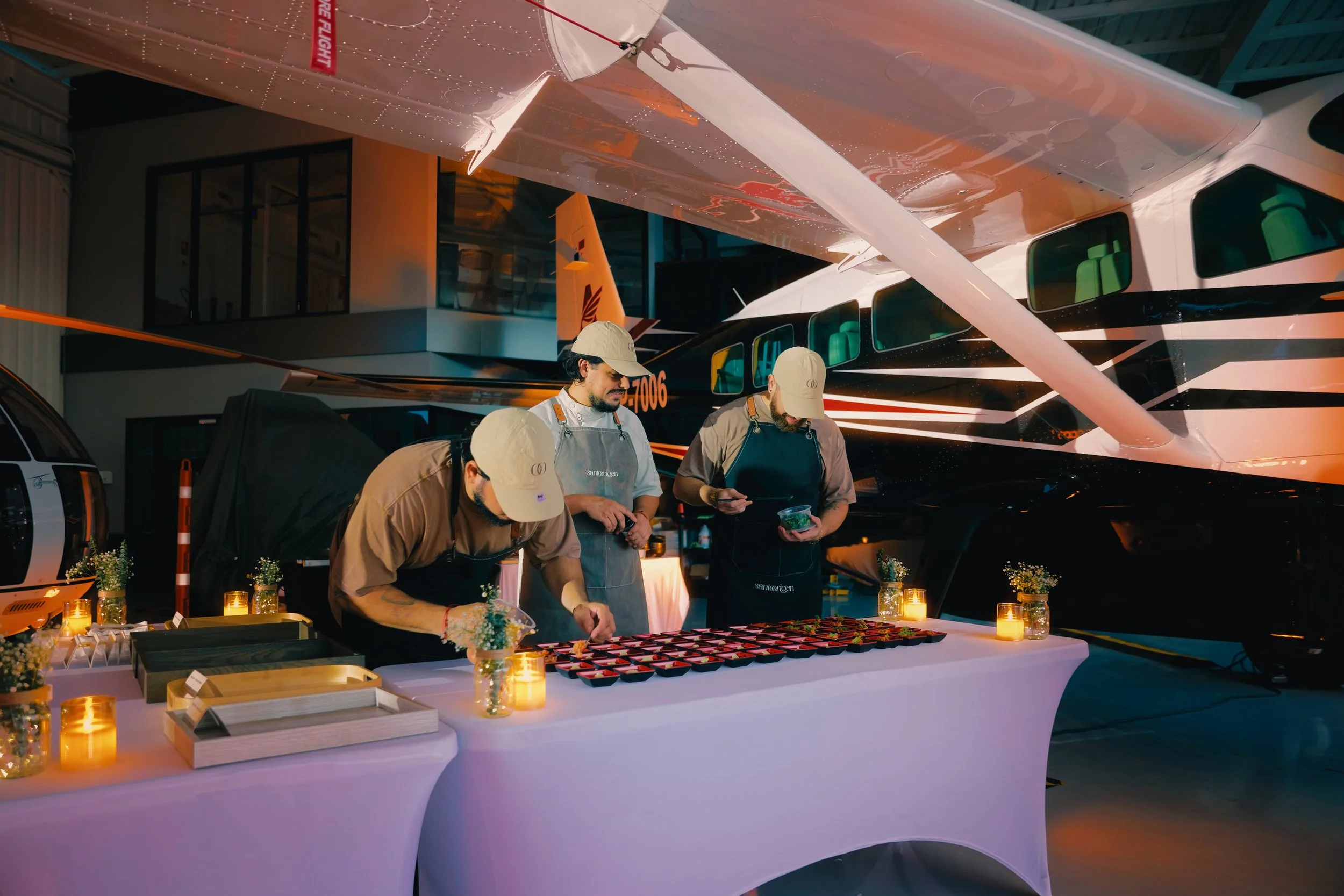 People preparing food or desserts at a table with flowers and candles inside an aircraft hangar, with a small airplane visible in the background.