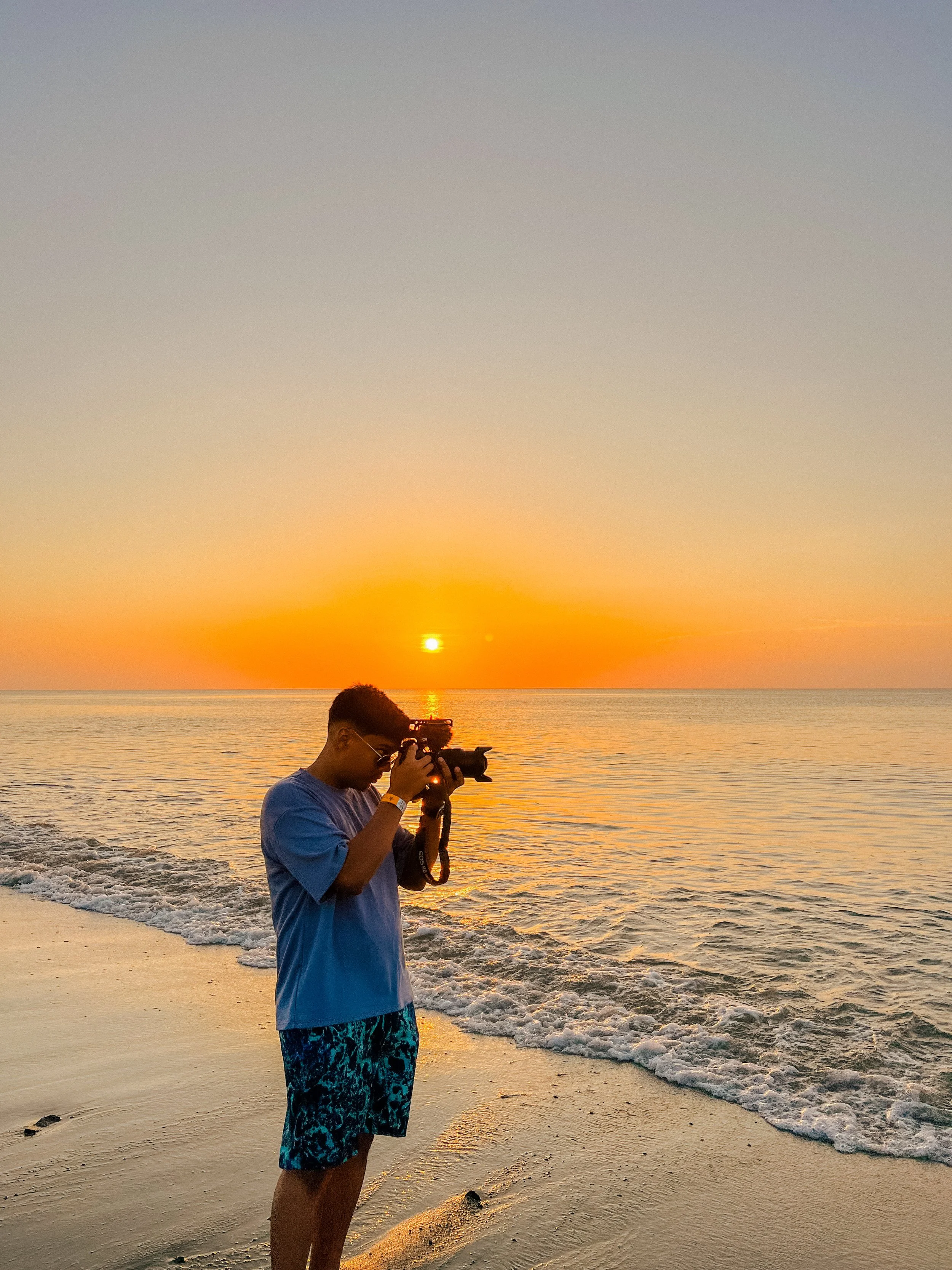 A young man is standing on a sandy beach at sunset, holding a camera and taking a photograph. The sun is low on the horizon over the ocean, casting a warm glow.