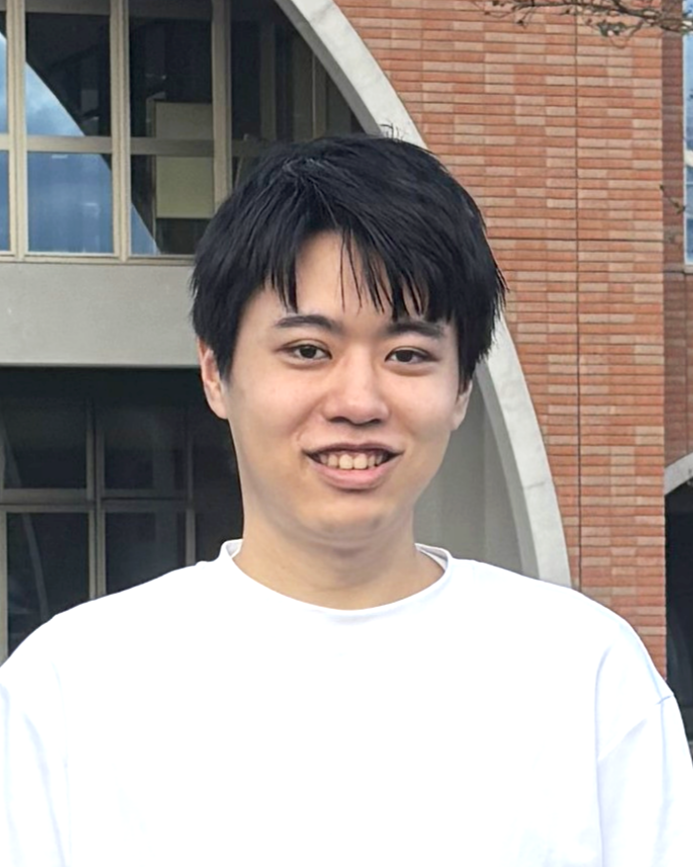 A young man with black hair smiling, wearing a white shirt, standing outside in front of a modern building with brick and concrete architecture.