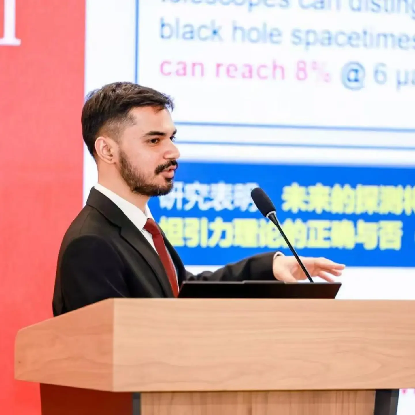 A man with dark hair and a beard is giving a presentation at a wooden podium with a microphone. There is a large screen behind him displaying a presentation slide with text in Japanese and English.