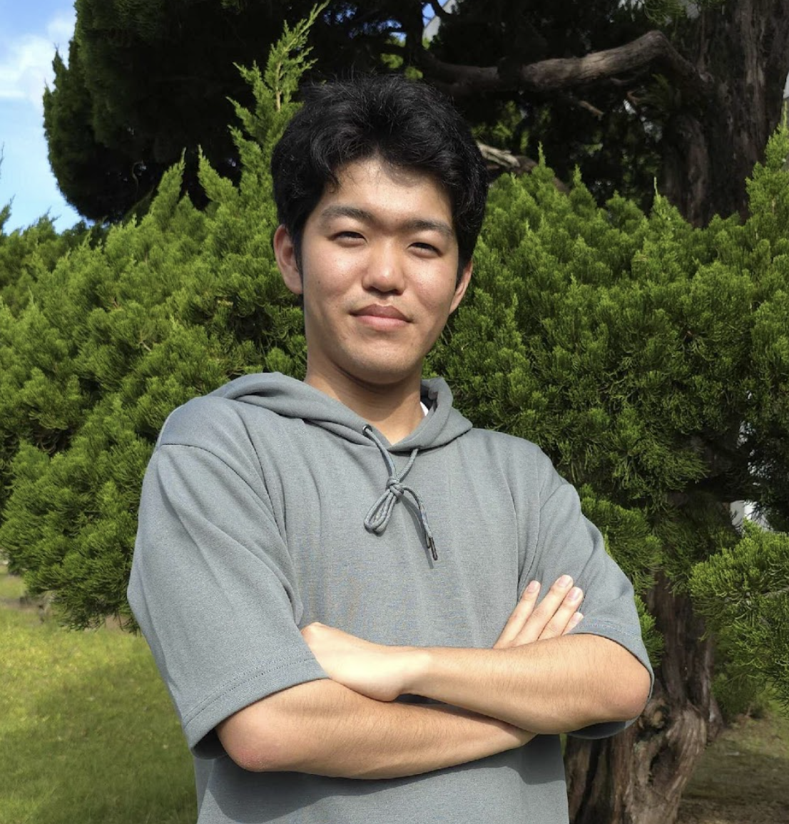 A young man with black hair wearing a gray hoodie standing outdoors in front of green trees and bushes.