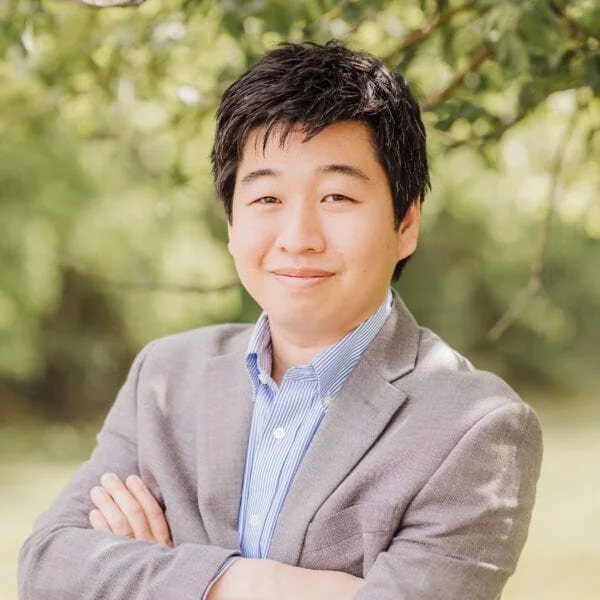 A young man with dark hair in a gray blazer and blue shirt, smiling with arms crossed outdoors, blurred green trees in the background.