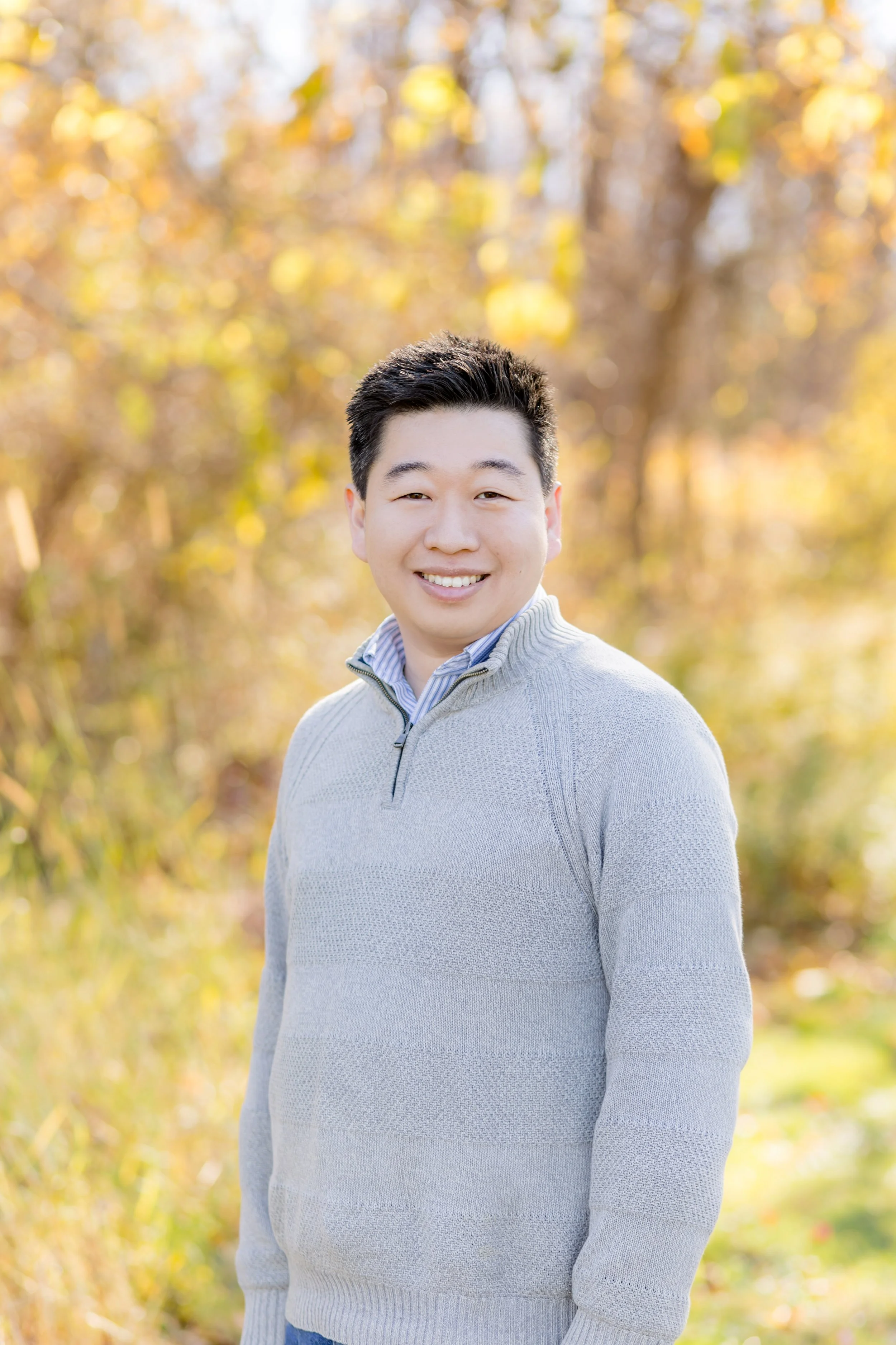 A young man with short black hair smiling outdoors in autumn with blurred yellow and orange fall foliage in the background.