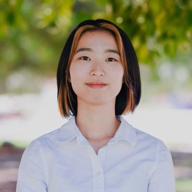 Young woman with shoulder-length hair and a white blouse outdoors, smiling with a blurred green background.