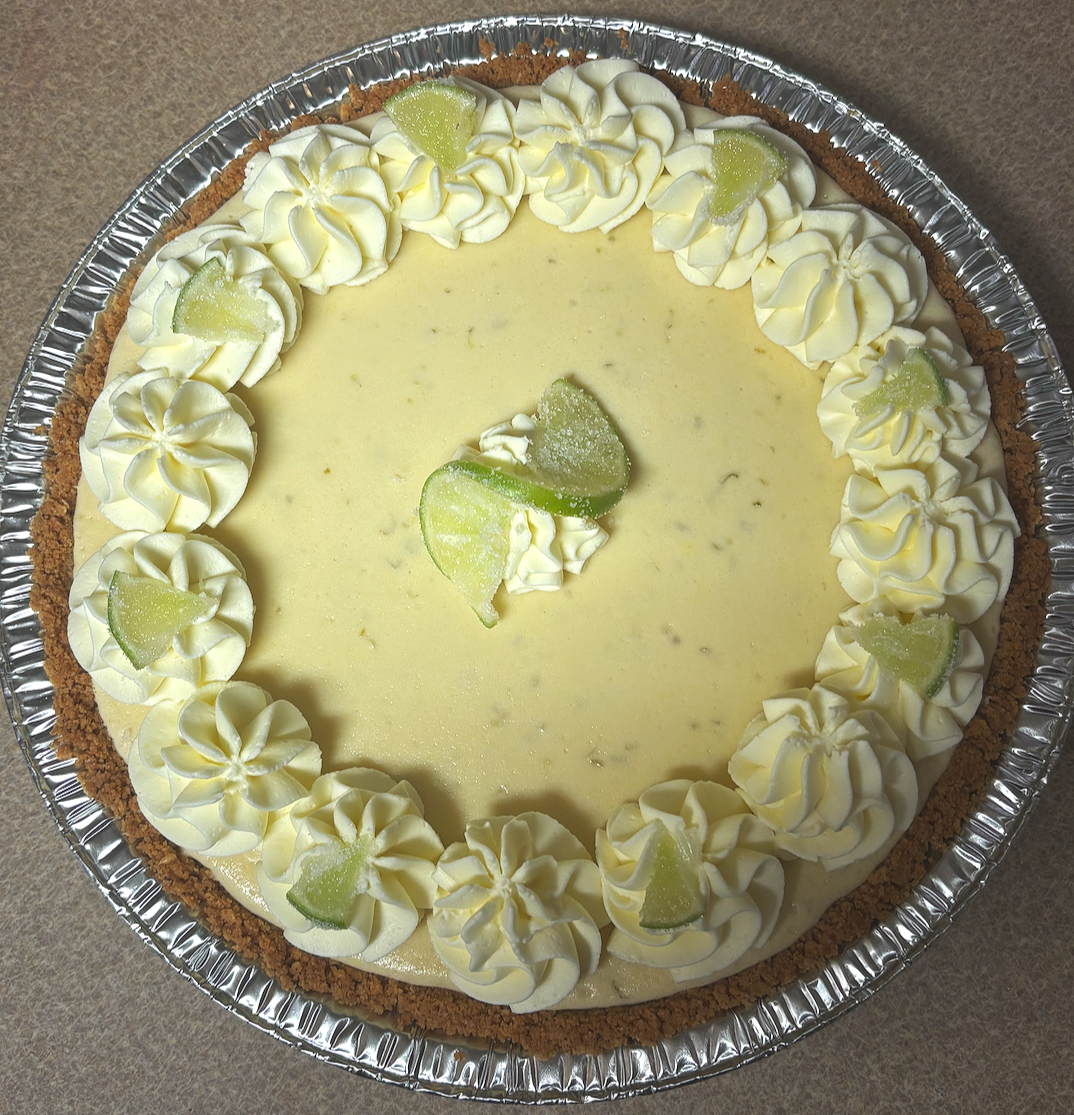 A lemon cheesecake with a graham cracker crust, decorated with whipped cream and slices of lime on top, in a round aluminum container.