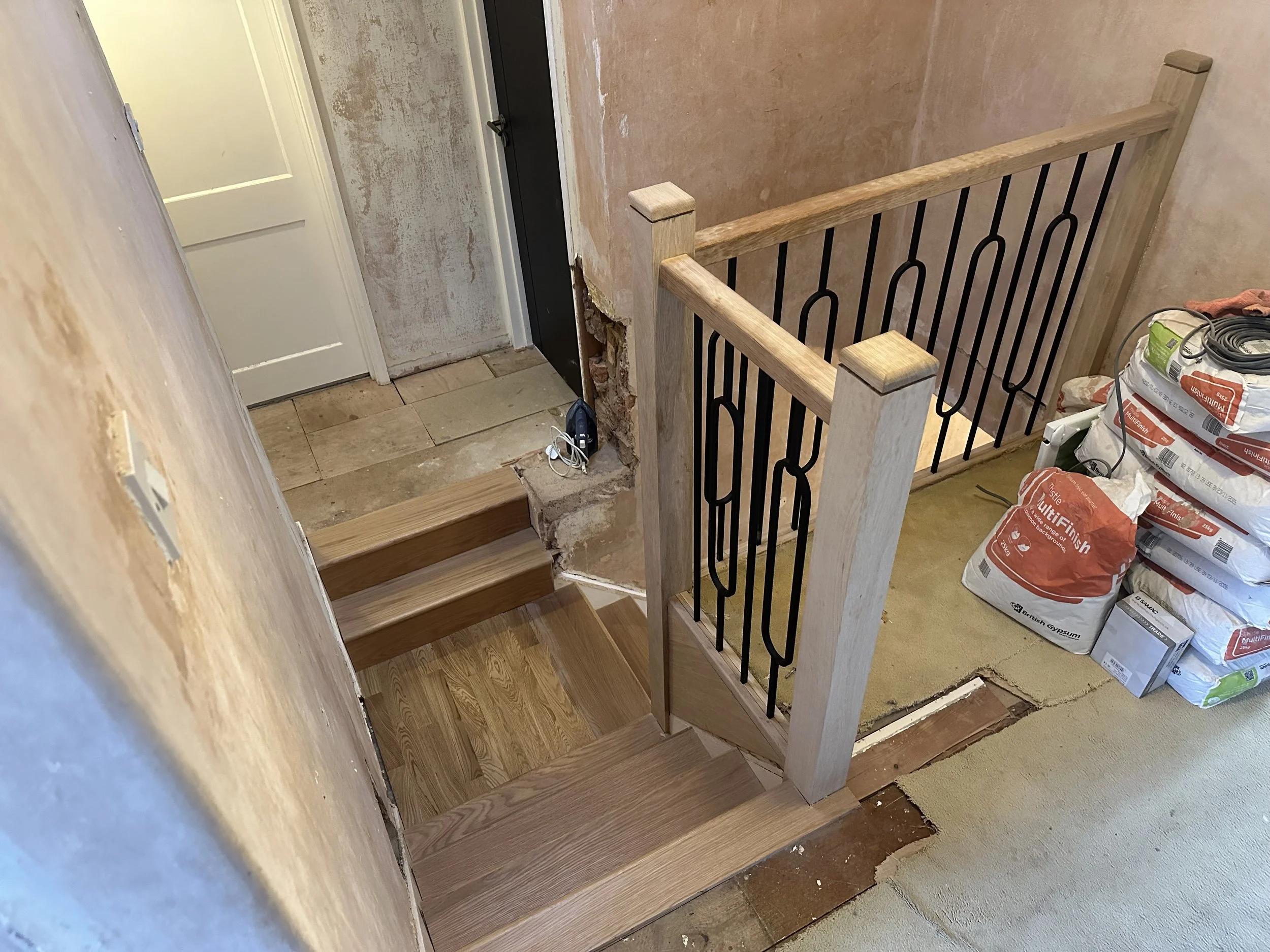 Interior staircase area under construction with wooden steps, a partially built railing, bags of construction materials, and exposed wall surfaces.
