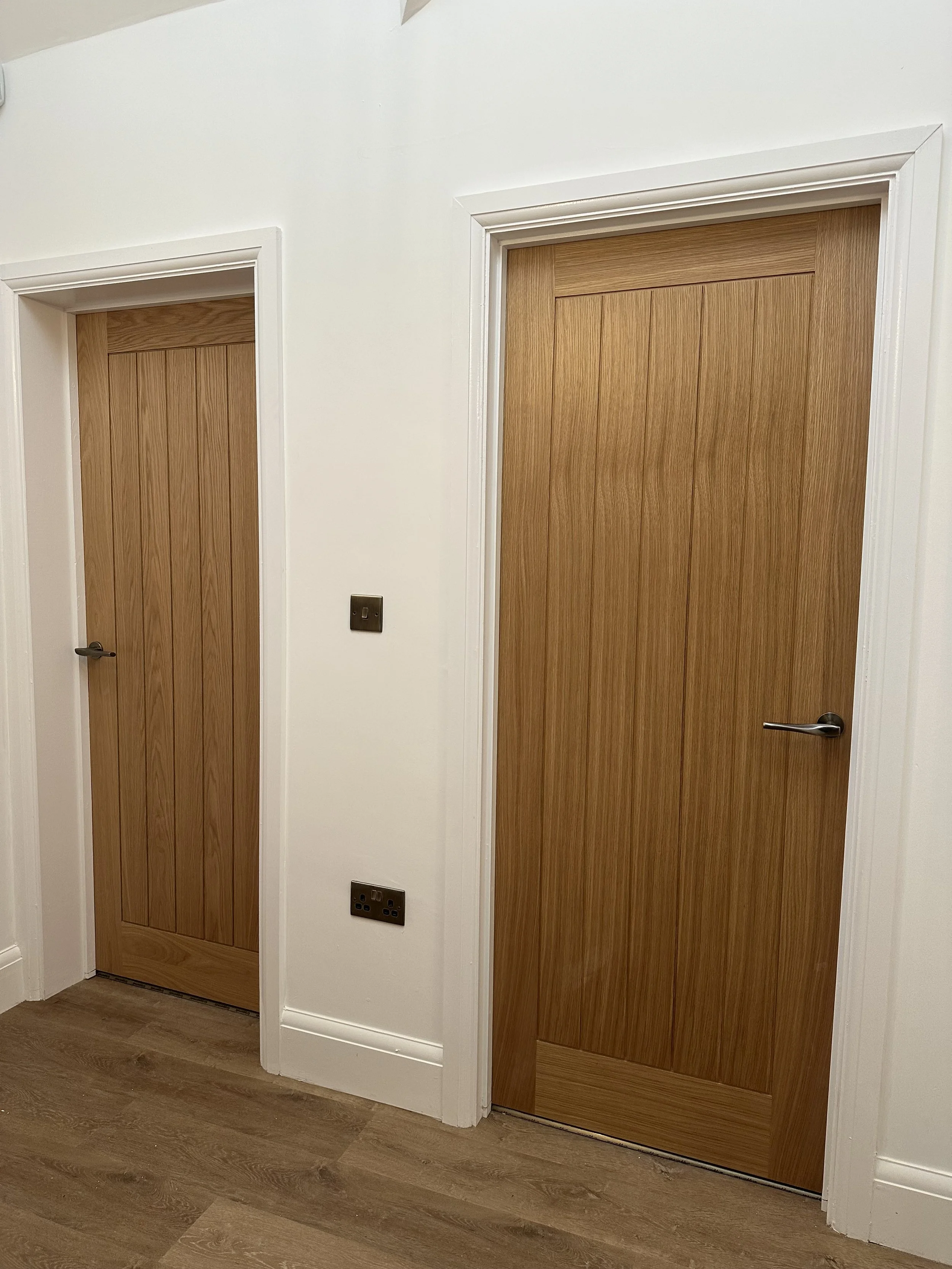 Two wooden doors with white trim in a hallway with wood flooring. One door is larger and has a modern handle; the smaller door has a black handle.