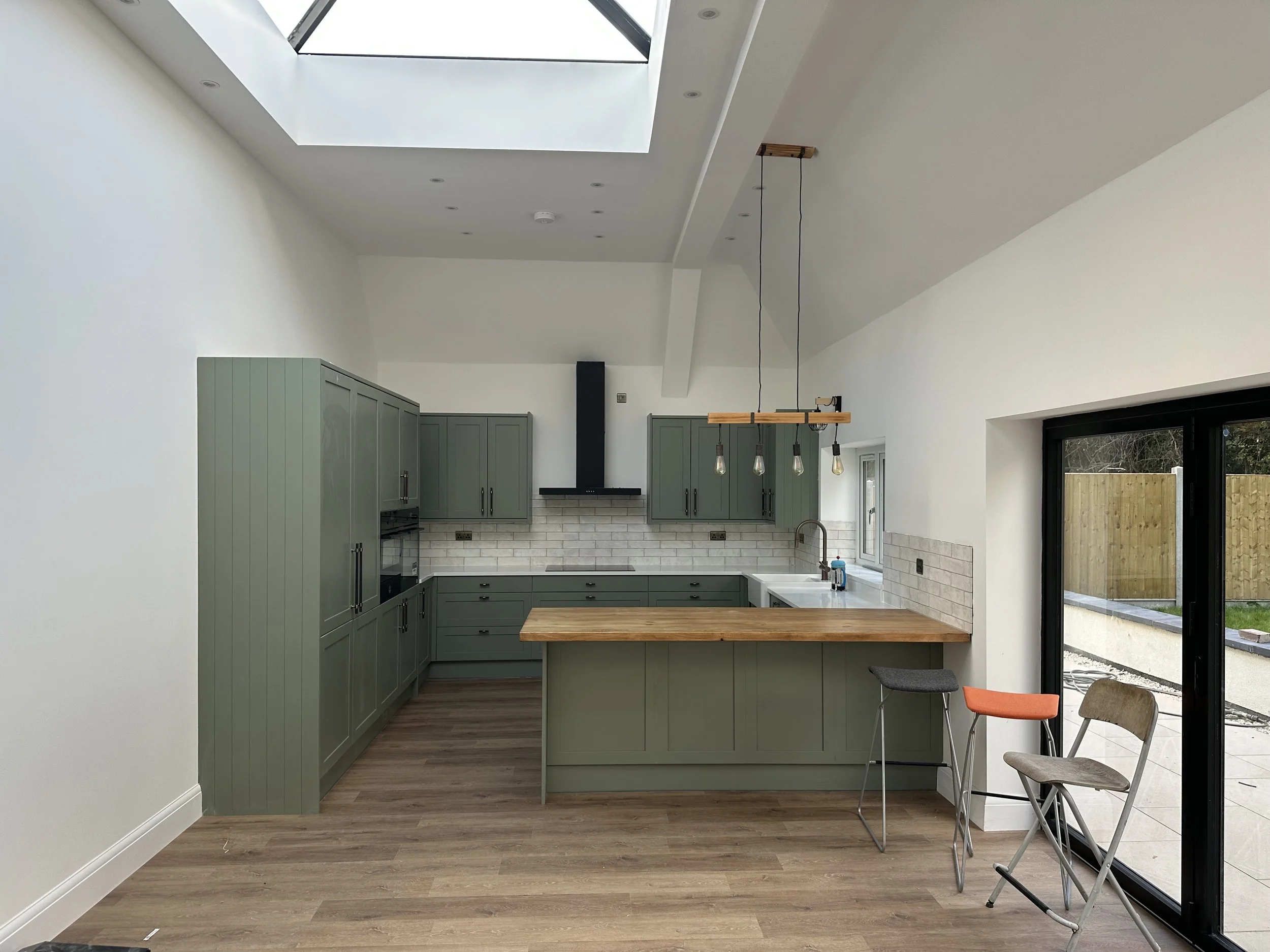 Modern kitchen with green cabinets, white brick backsplash, large skylight, and sliding glass door leading outside. There are three chairs with different colored seats near the sliding door, and a wooden breakfast bar.