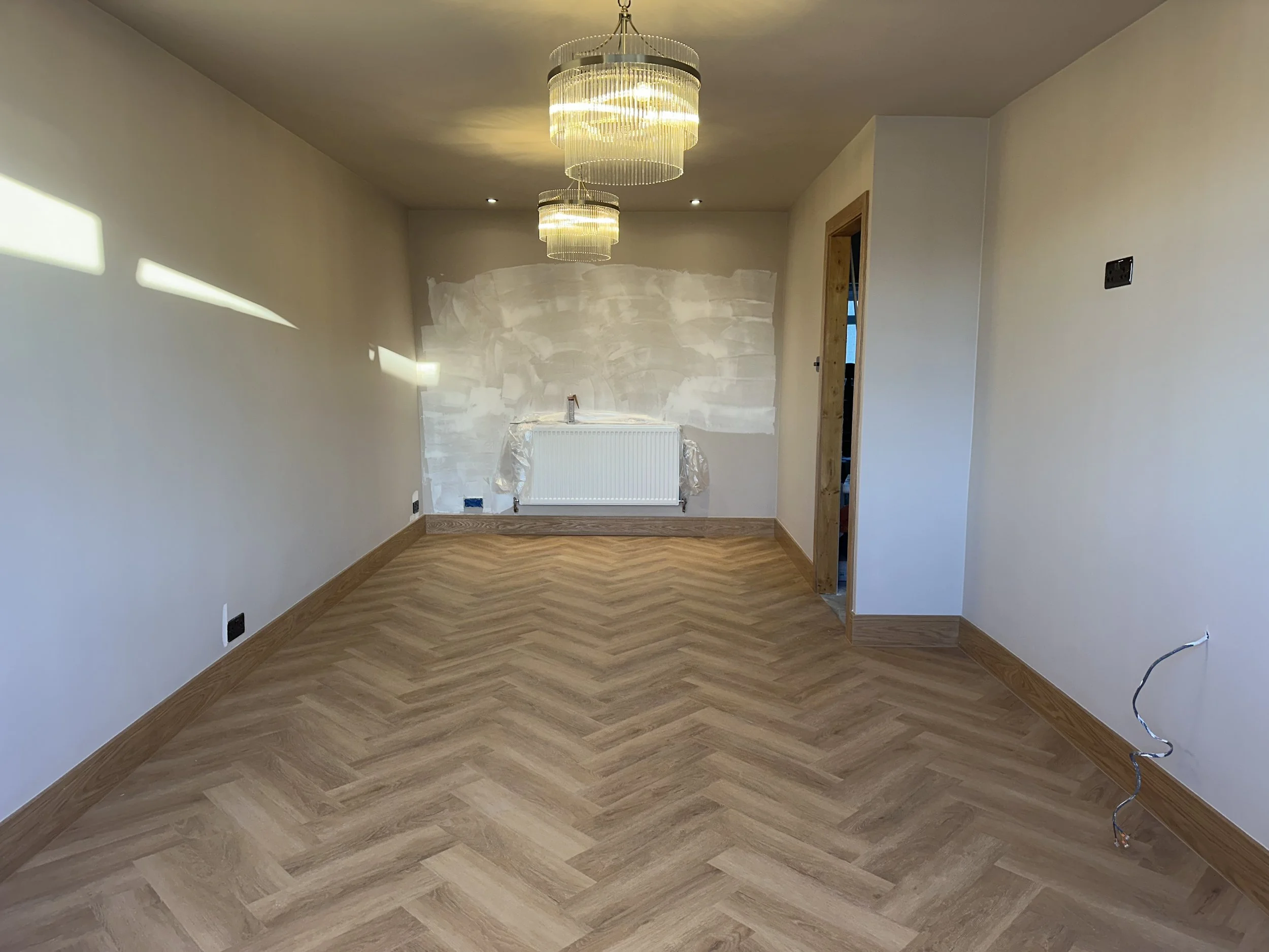 Empty room with beige walls, wood flooring, two modern chandeliers hanging from the ceiling, a radiator under construction with plastic covering, a door frame to the right, black electrical outlets and a plug cable on the walls, and sunlight casting shadows on the left wall.