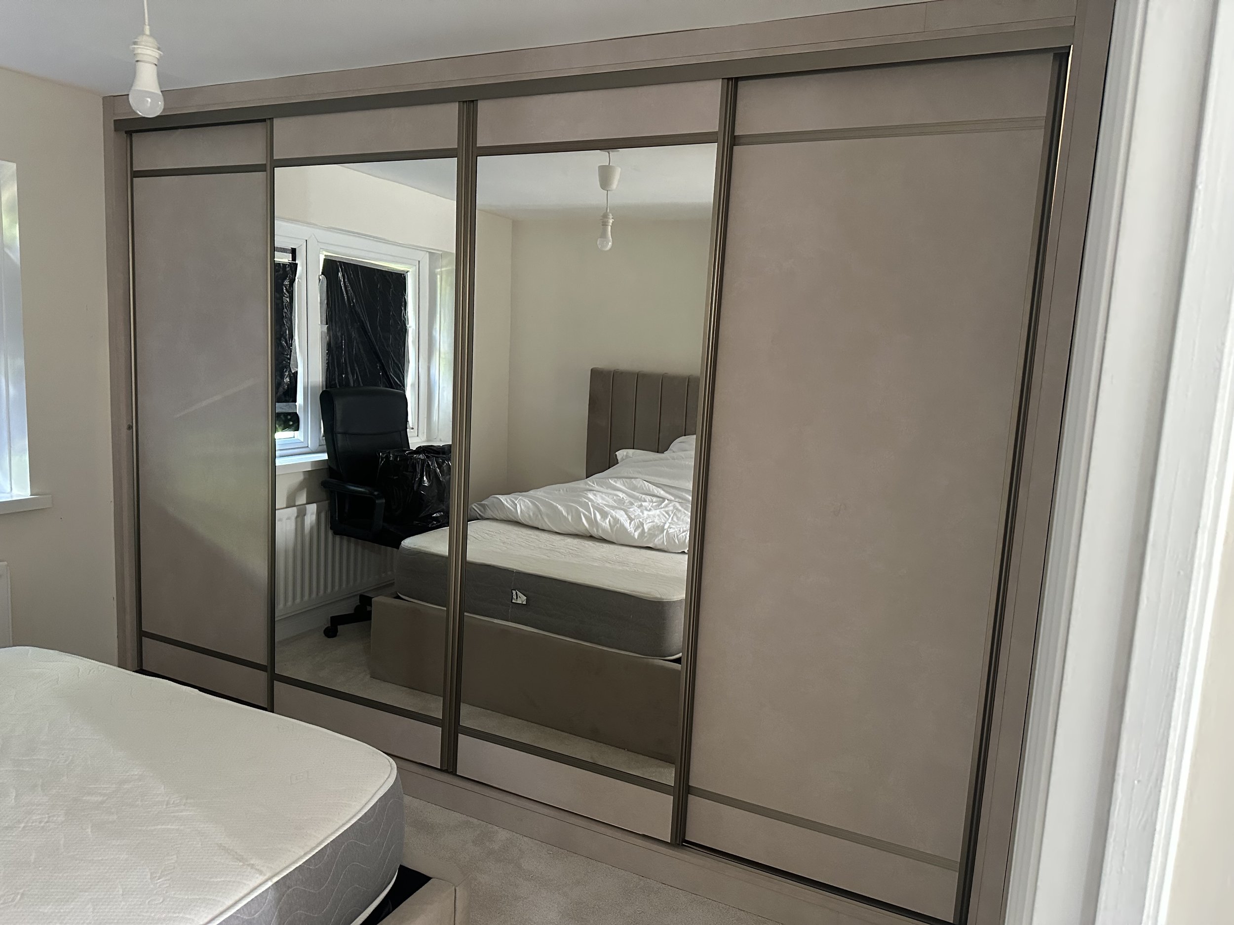 Bedroom with a mirrored sliding door wardrobe, a bed with white bedding, a window with blinds, a black office chair, and light fixtures hanging from the ceiling.