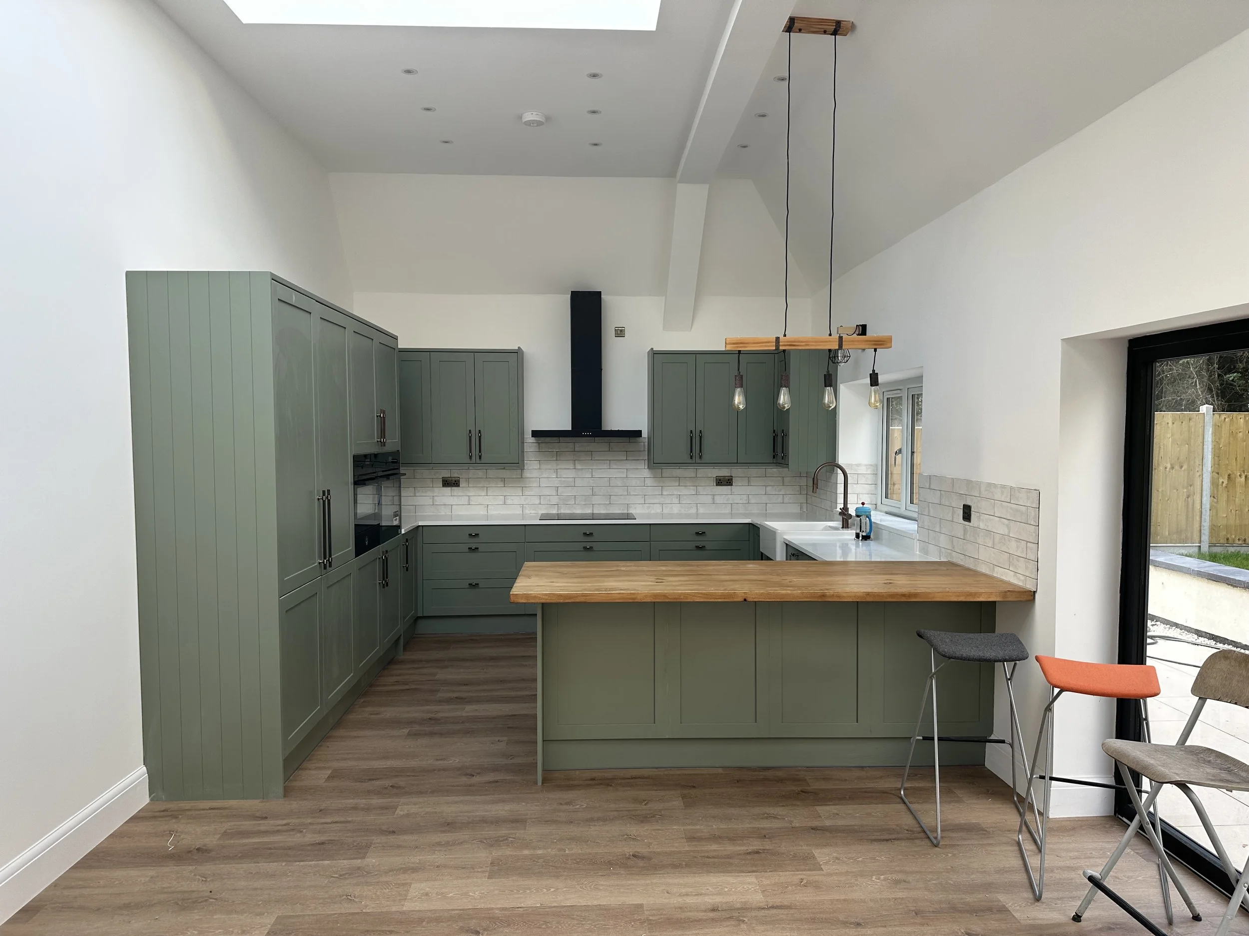 Modern kitchen with green cabinets, white countertop, brick backsplash, and hanging Edison-style light bulbs. Large window with door leading outside, wooden floor, and a small bar stool.