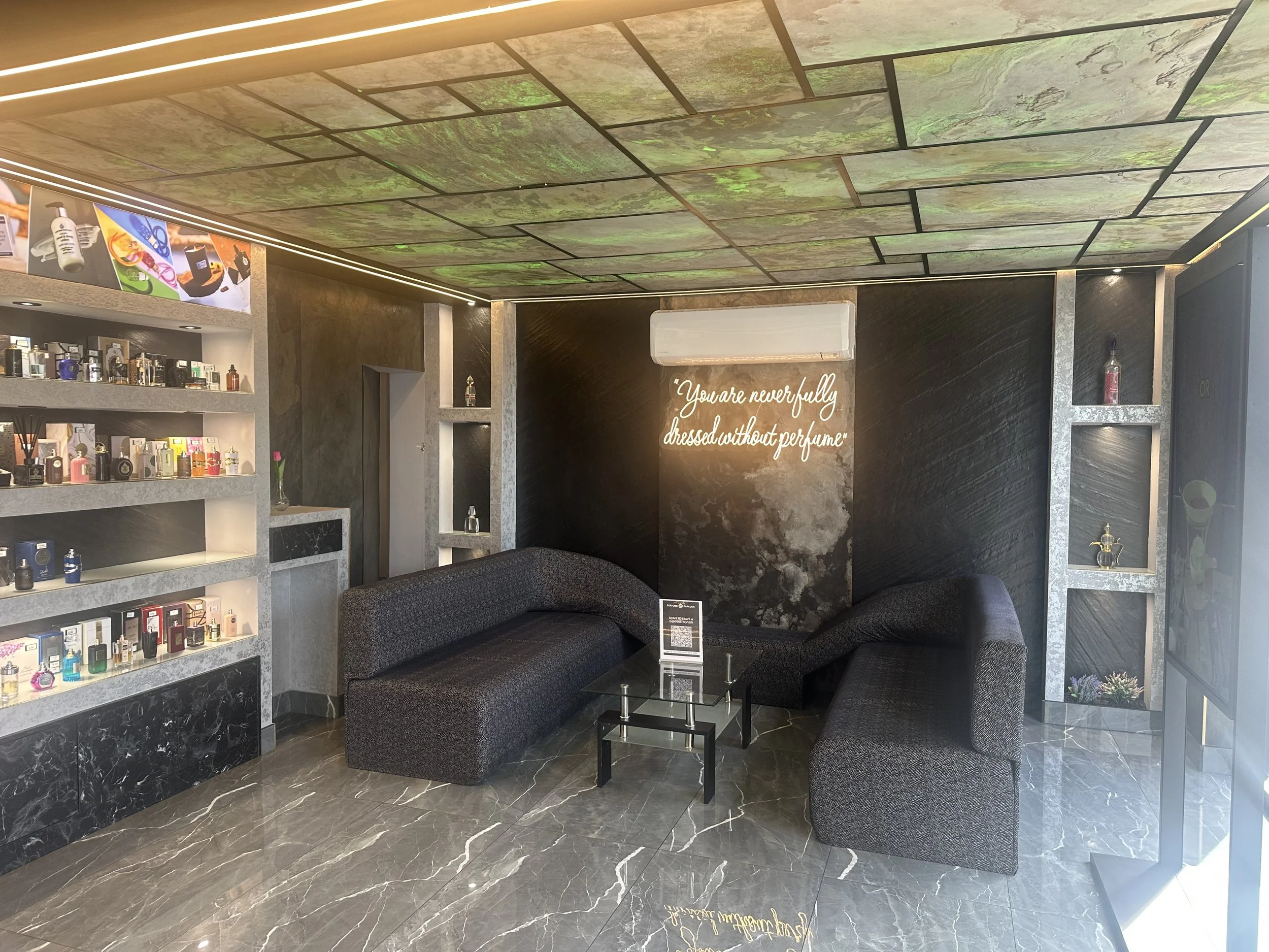 Modern interior room with marble flooring, black upholstered couches, glass coffee table, black textured wall, wall-mounted quote, display shelves with perfume bottles, decorative items, and a ceiling with greenish marble tiles.